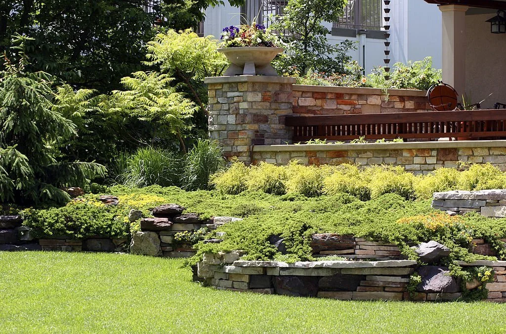 Tiered stone retaining walls with dense greenery provide structure and visual interest in a well-maintained garden setting.