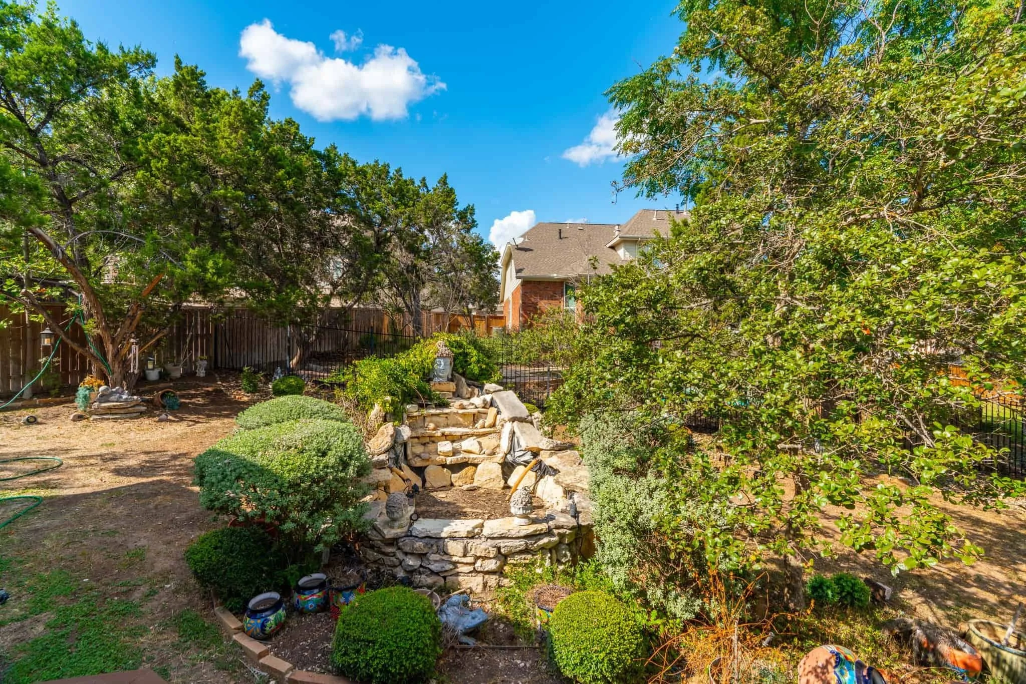 Natural backyard with mature trees, rocky waterfall feature, trimmed hedges, and rustic landscaping under a clear blue sky.