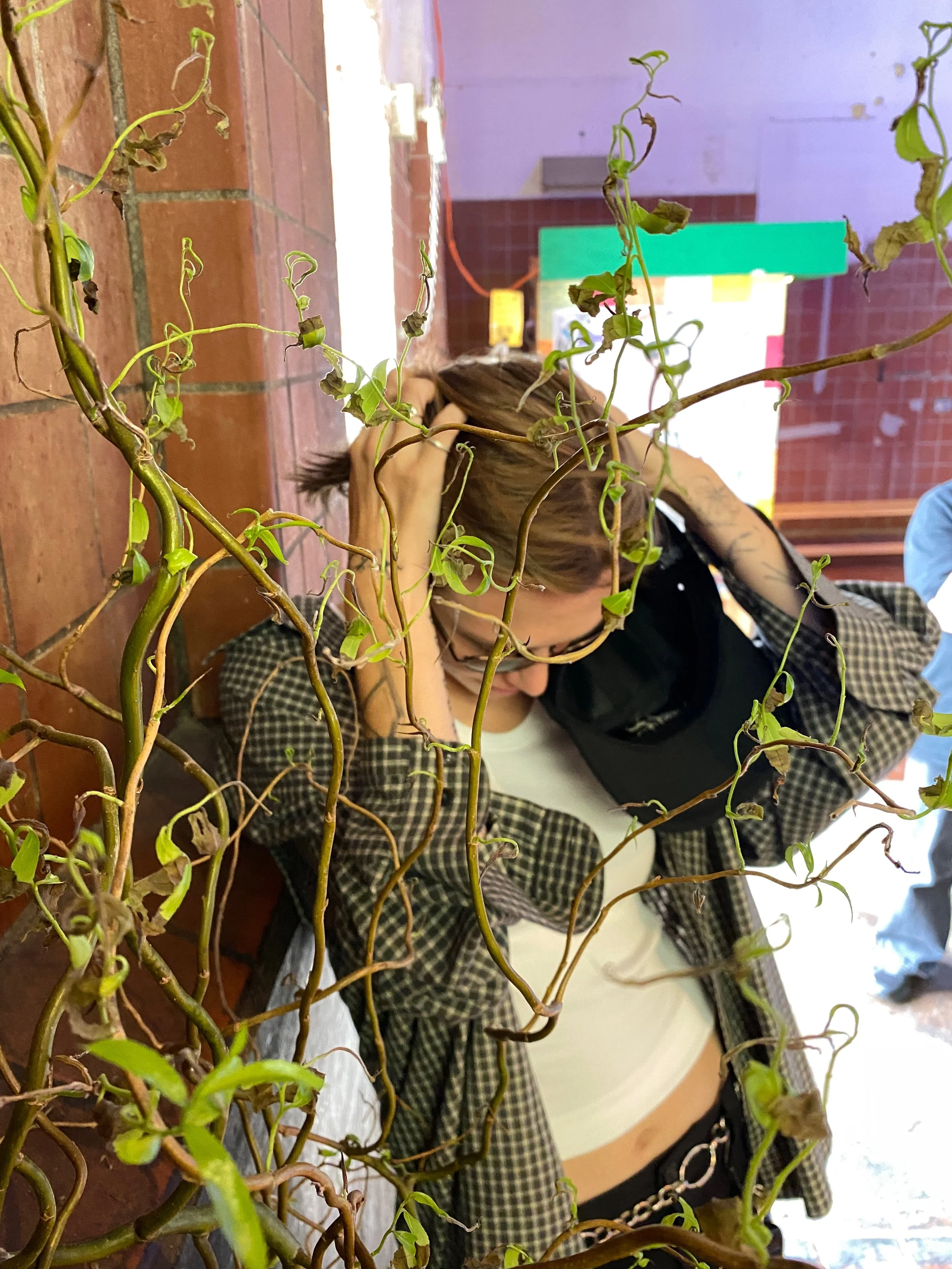 A woman leaning against a brick wall, partially obscured by a tangle of vine plants with green leaves.