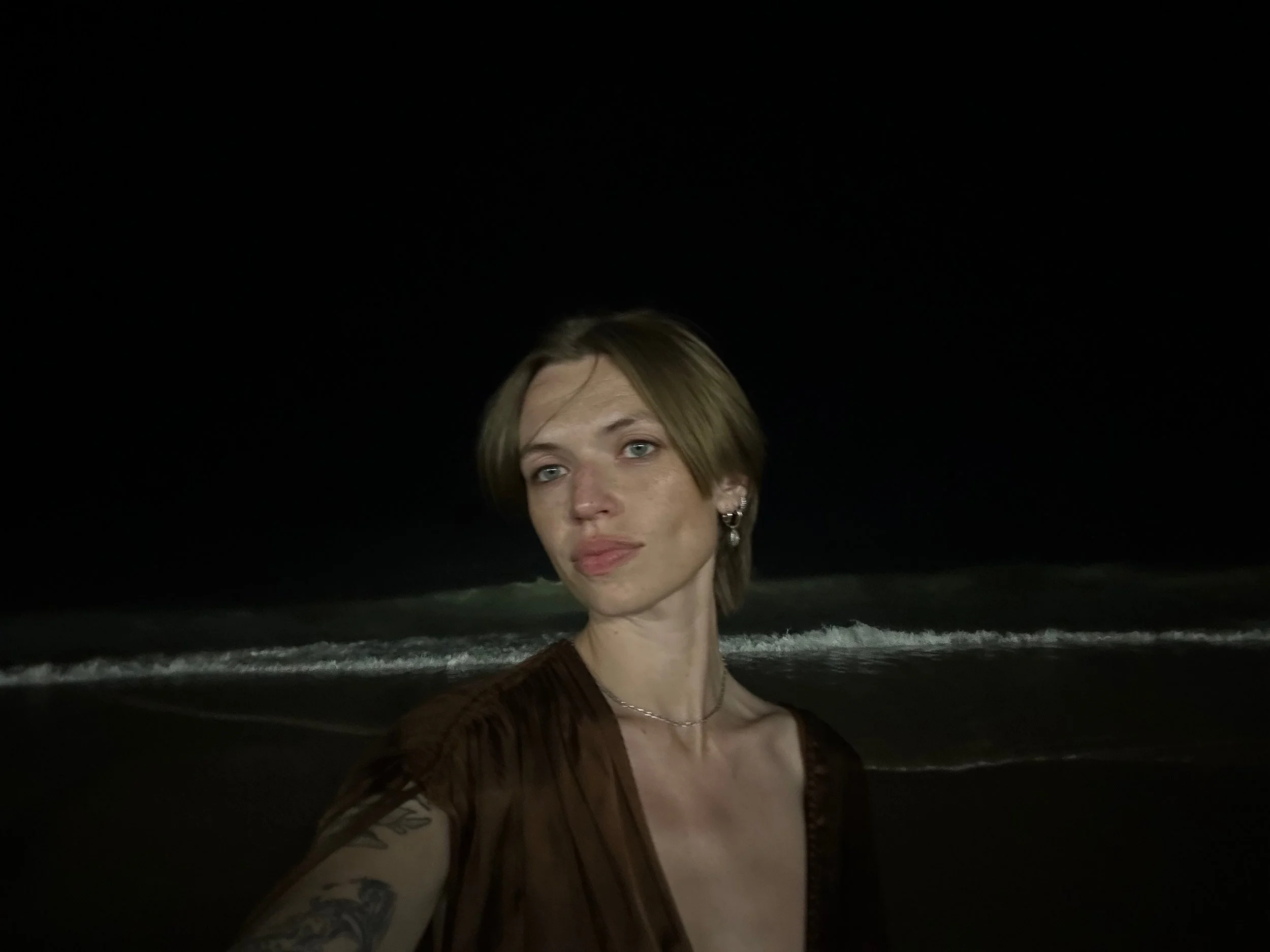 A woman taking a selfie at night on a beach, with dark sky and ocean waves in the background.