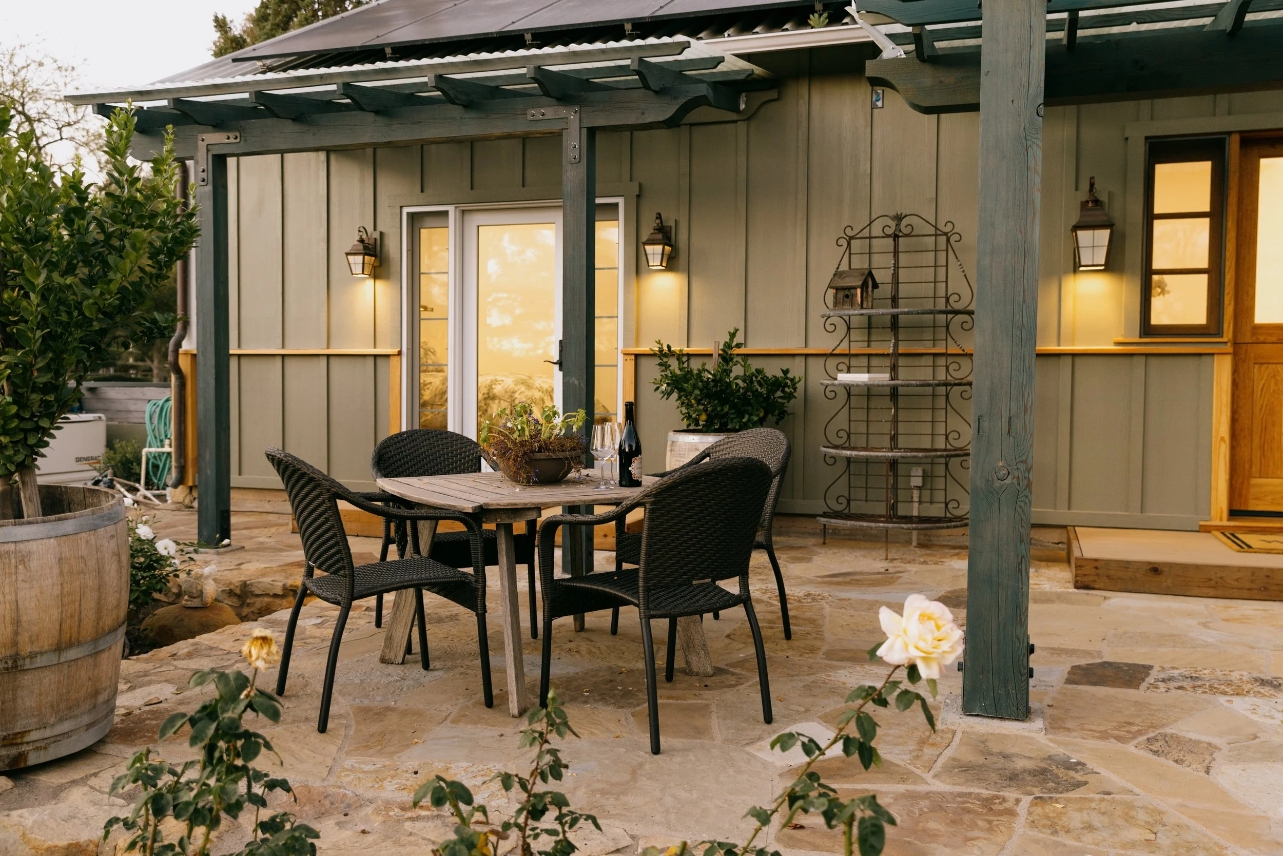 Outdoor patio with a wooden table, four black chairs, a bottle of wine, glasses, and plants, with a house in the background during sunset.