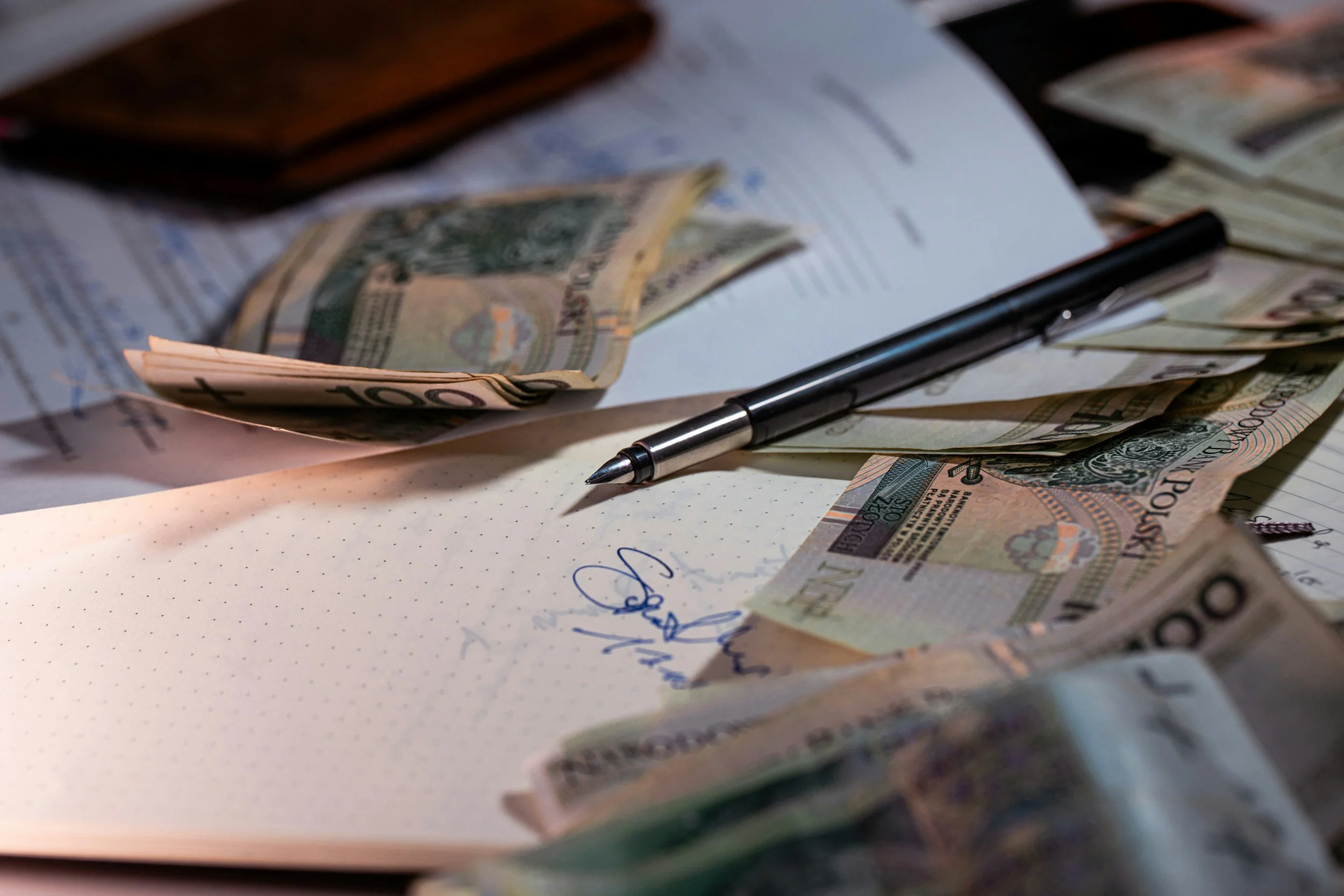 A black pen lying on an open dotted notebook with handwritten text, surrounded by scattered U.S. currency bills including $100 and $20 bills, on a cluttered desk.