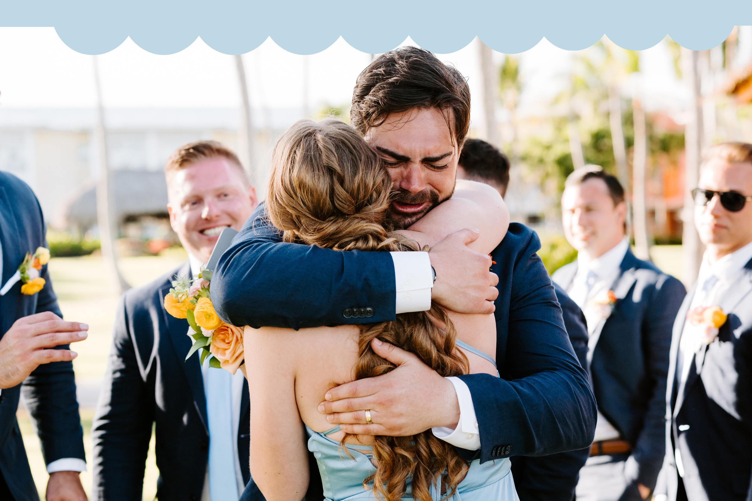 A man in a dark suit and a woman in a dress are hugging tightly, showing emotions. Several men in suits stand in the background, smiling and watching the scene. It appears to be an outdoor wedding or celebration with a festive and emotional atmospher