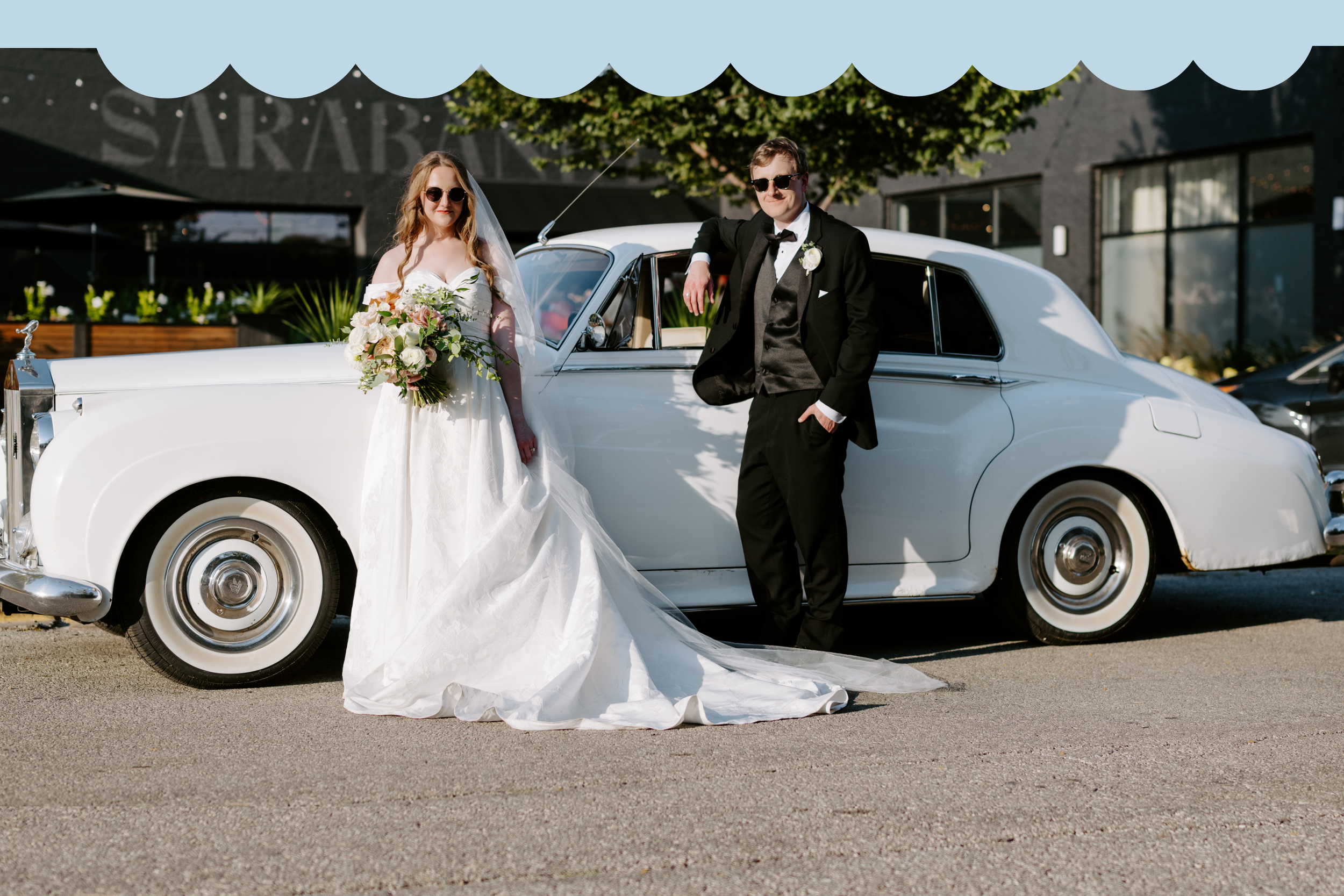 A bride in a white wedding gown holding a bouquet of flowers and a groom in a black tuxedo with sunglasses standing next to a vintage white car