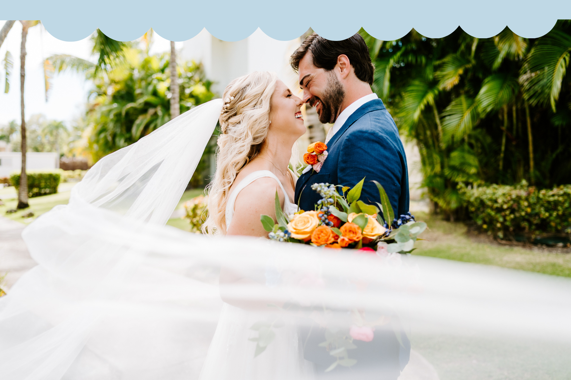 A bride and groom are outside, smiling and touching foreheads, with the bride holding a bouquet of orange roses and greenery, and her veil flowing in the wind. Tropical plants and trees are in the background.