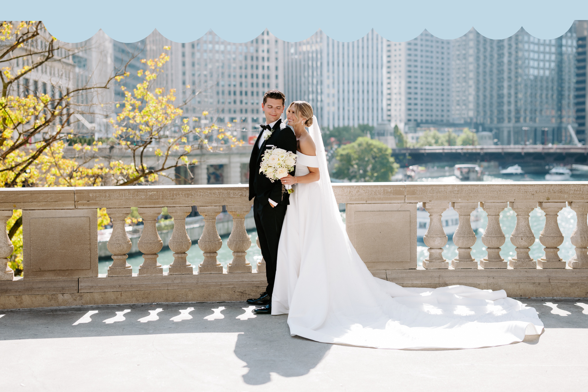 A bride and groom dressed in wedding attire standing on a stone balcony with a cityscape and waterfront in the background. The bride is holding a bouquet of flowers, and they are smiling at each other.