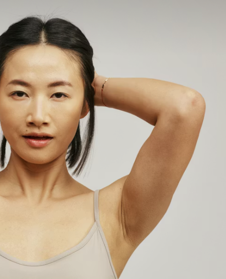 A woman with black hair in a ponytail, wearing a light-colored tank top, raising her arm and touching her head against a plain white background.
