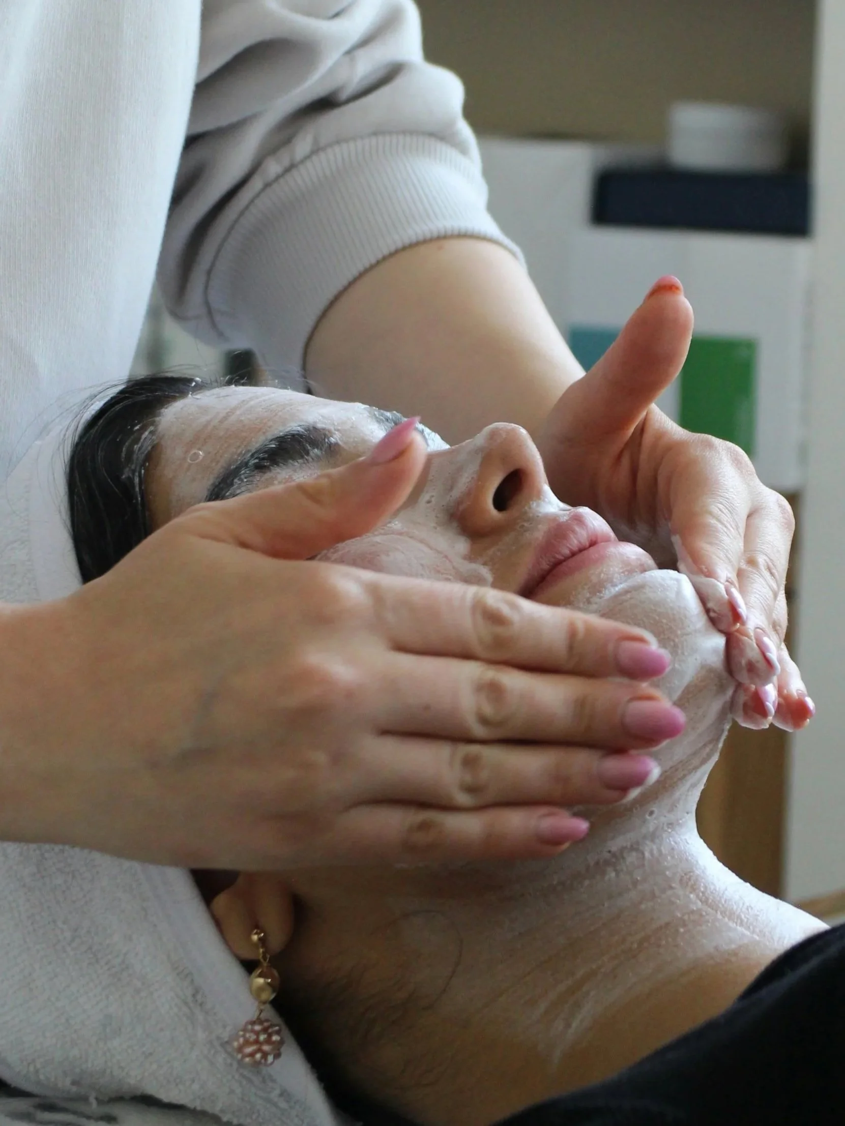 A woman receives a facial treatment from a professional esthetician, who is applying a facial mask or cleanser while gently massaging her face.