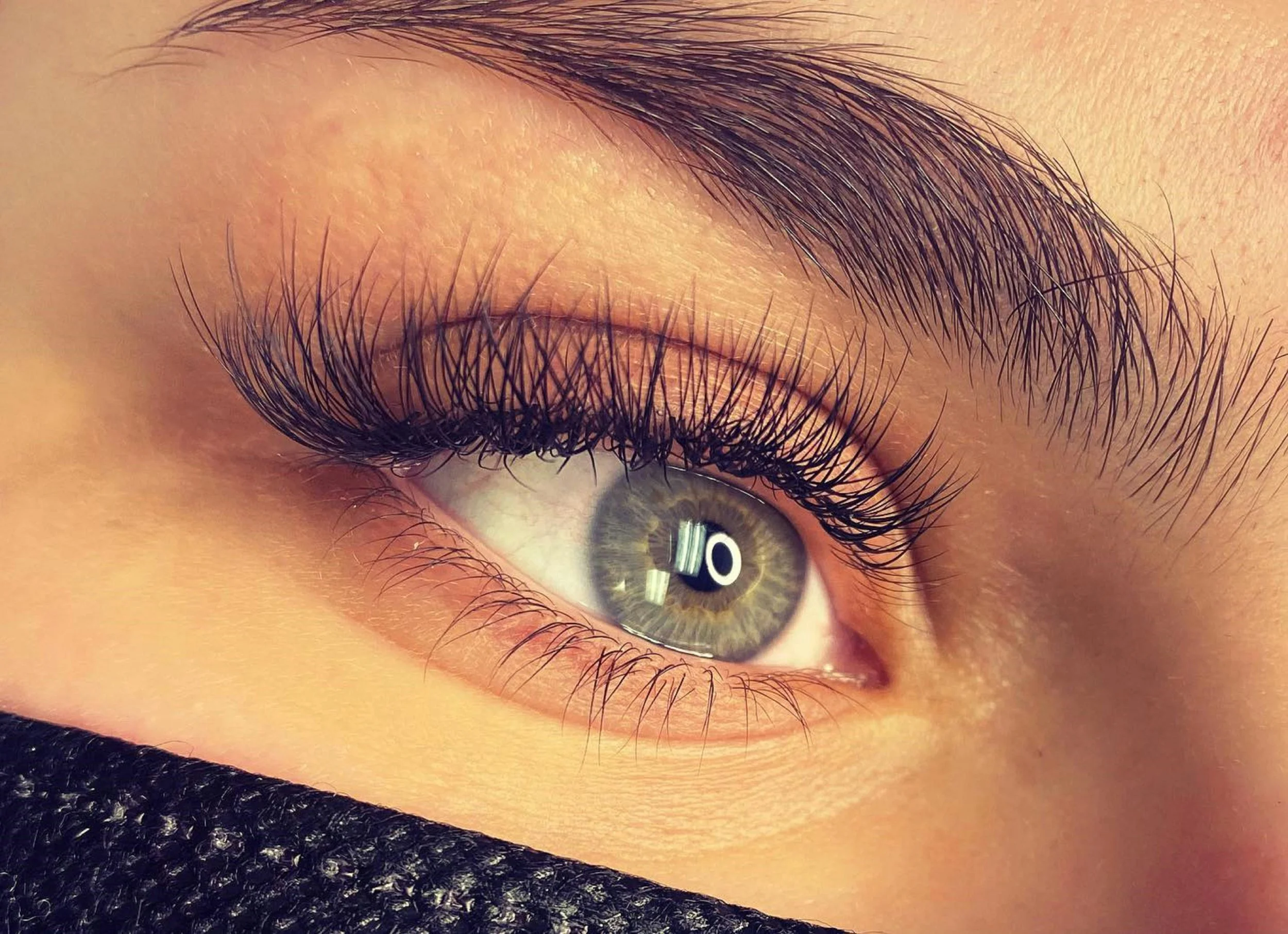 Close-up of a human eye with hazel iris, long eyelashes, and well-defined eyebrows, with a reflection of a ring light in the eye.