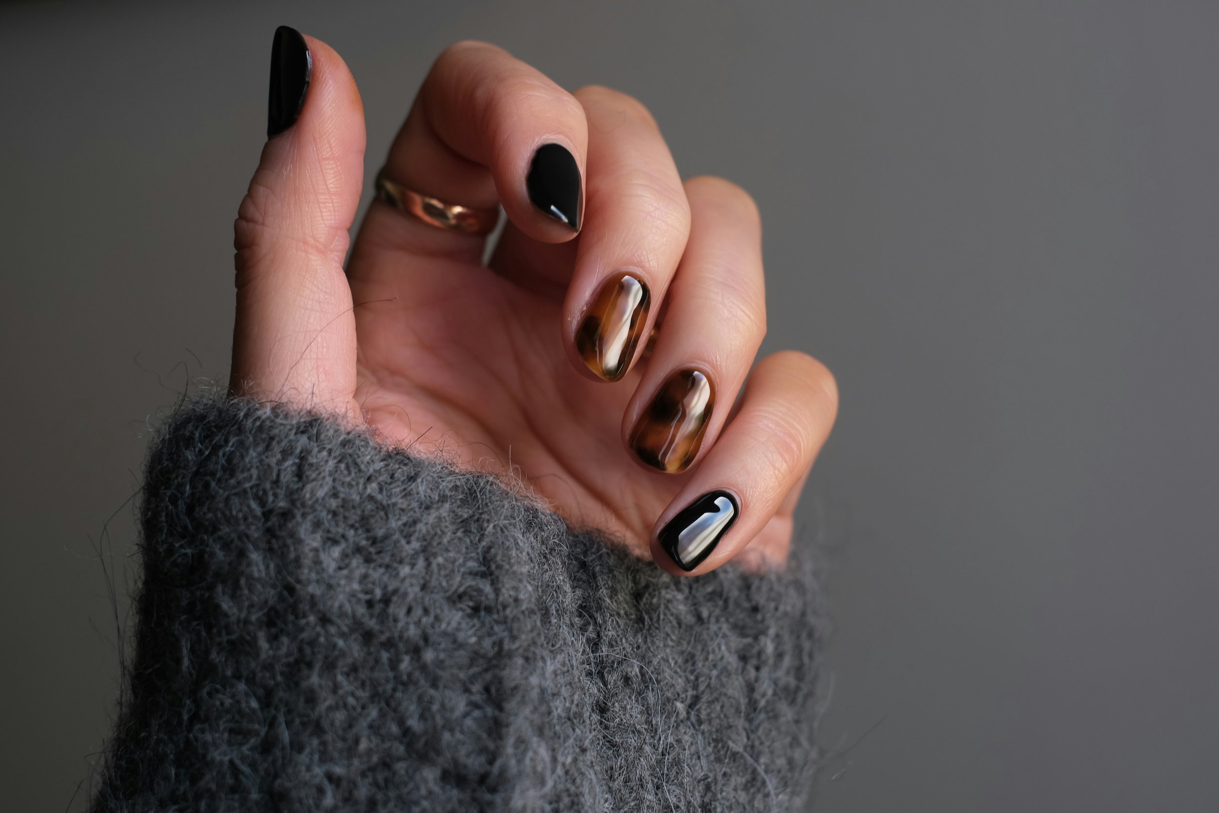 Close-up of a hand with manicured nails featuring black, tortoiseshell, and black with white accents, wearing a gray fuzzy sleeve against a gray background.