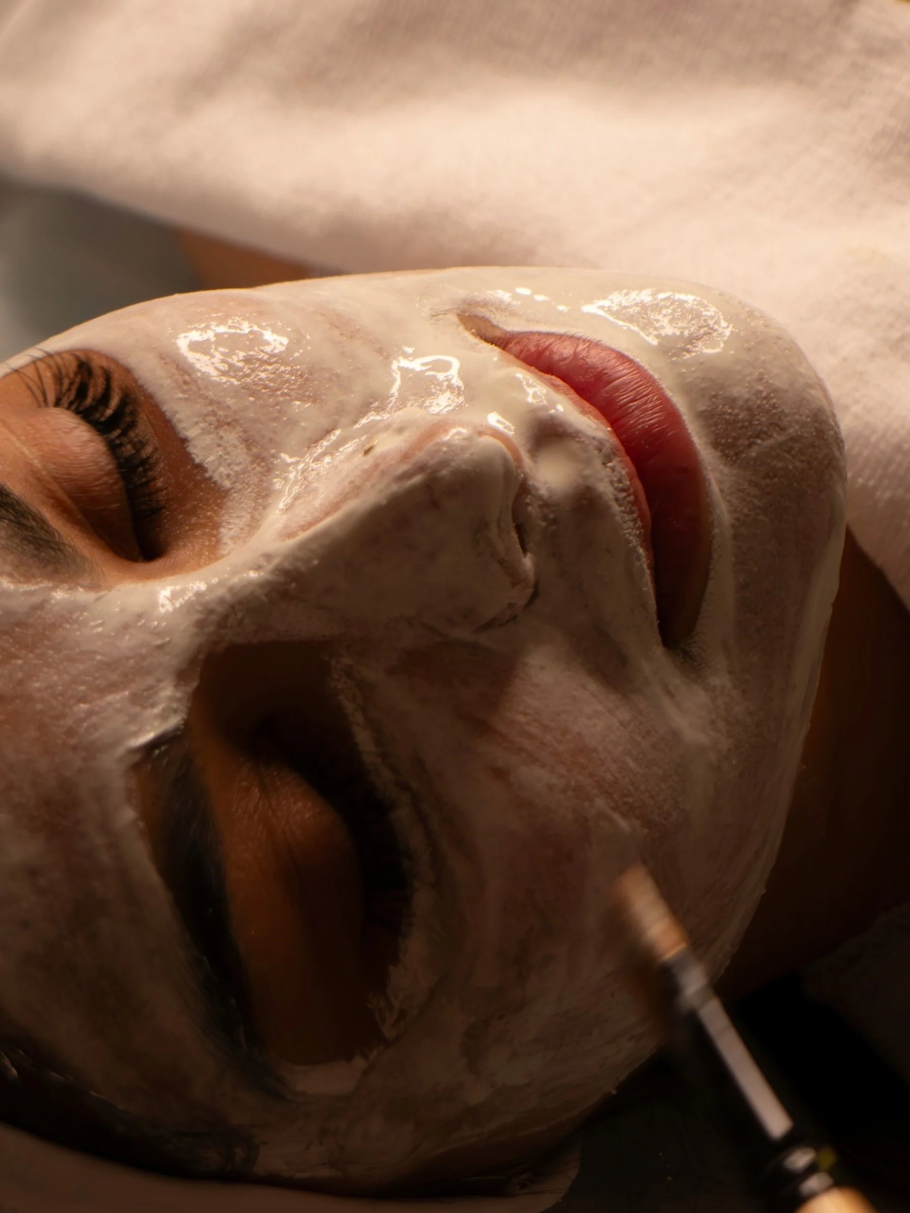 A woman lies down with her eyes closed, receiving a facial treatment with a white mask applied to her face, while a professional uses a small tool on her chin.