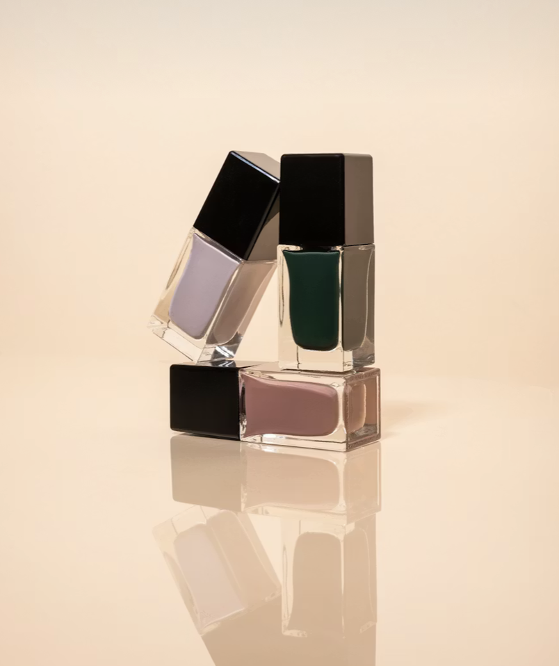 Three bottles of nail polish, with one leaning on the other two, reflected on a smooth surface with a soft beige background.