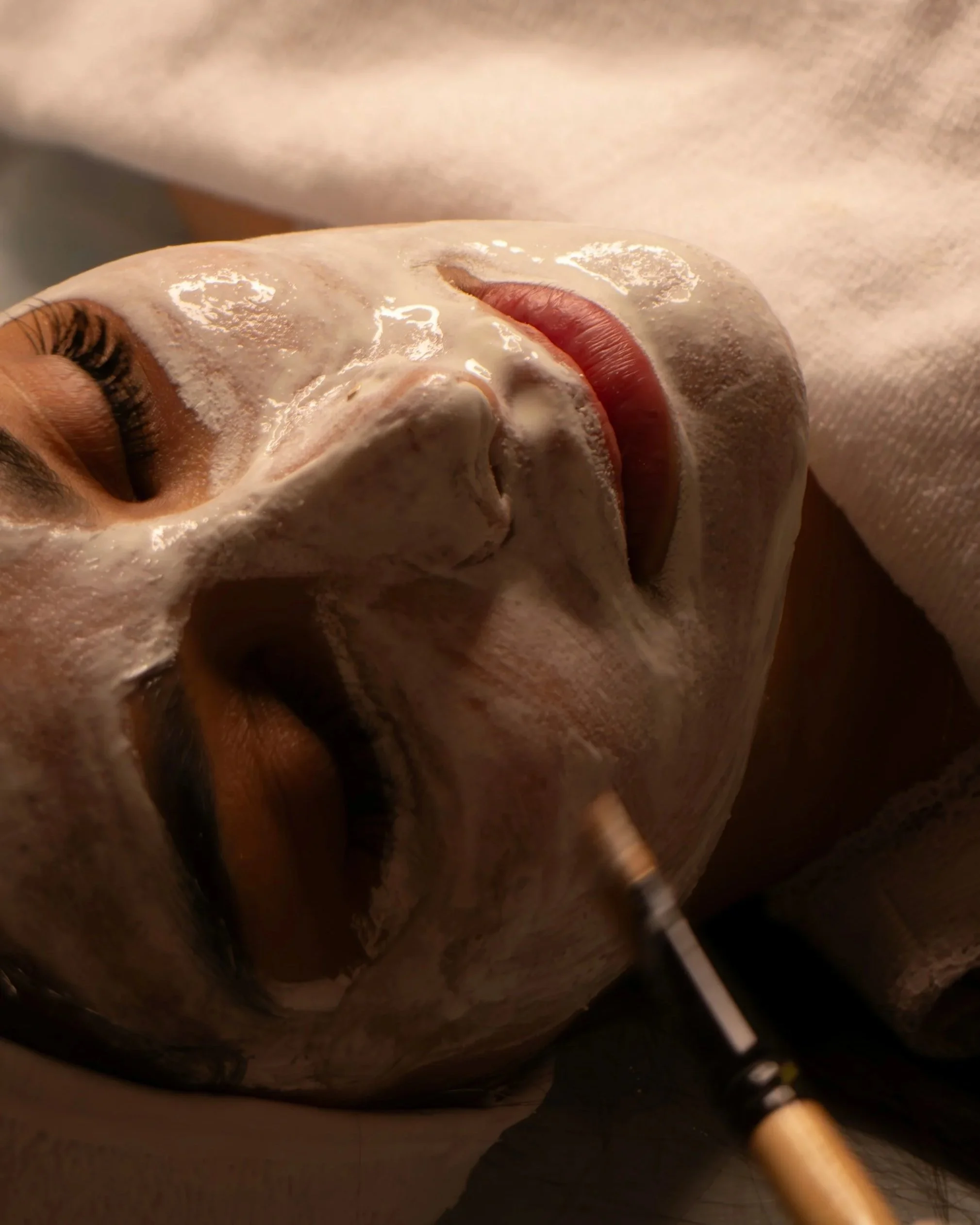 A person with closed eyes receiving a facial treatment, with a mask being applied to their face using a brush.
