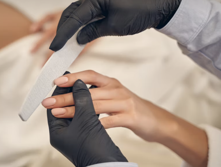 A nail technician applying nail polish to a woman's fingernails in a salon, with the woman lying on a bed.