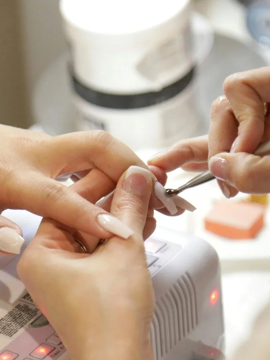 Close-up of a person's hands receiving a manicure, with a nail technician applying clear top coat on the nails using a brush.