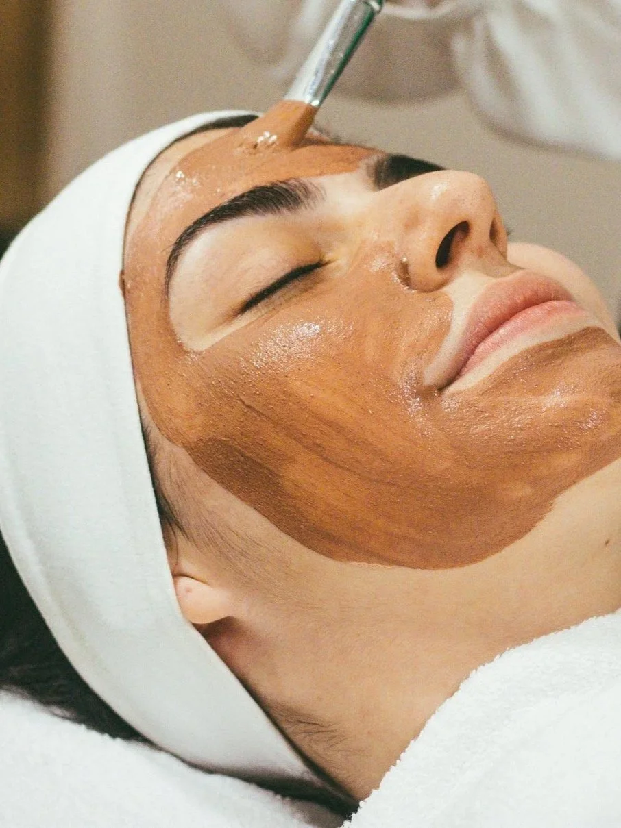 Woman receiving a facial treatment, with a brown mask applied to her face while lying down with eyes closed, wearing a white headband.