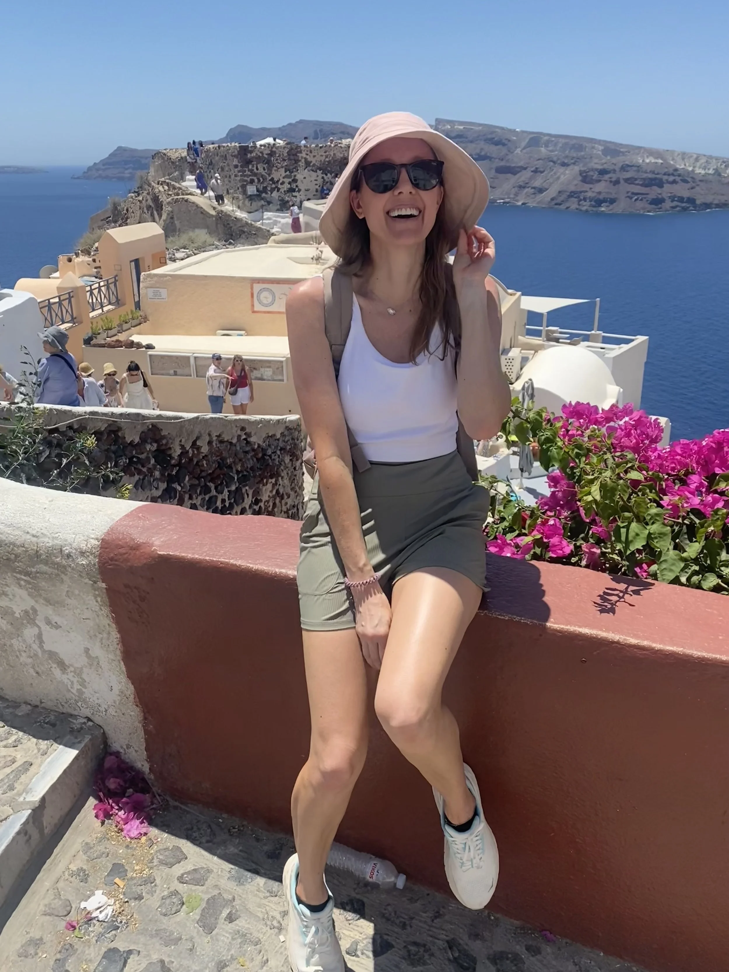 A woman smiling, wearing sunglasses, a wide-brimmed hat, a white sleeveless top, green shorts, and white sneakers, leaning against a red wall with pink flowers, overlooking a blue sea and a cliffside town.