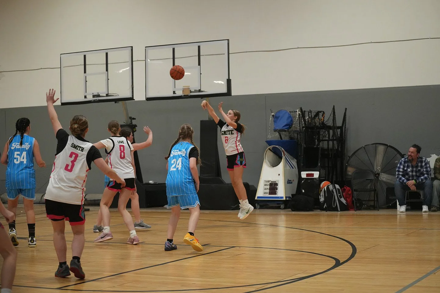 Girls playing basketball in an indoor gym, one girl is jumping and shooting the ball, others are watching or preparing to defend.