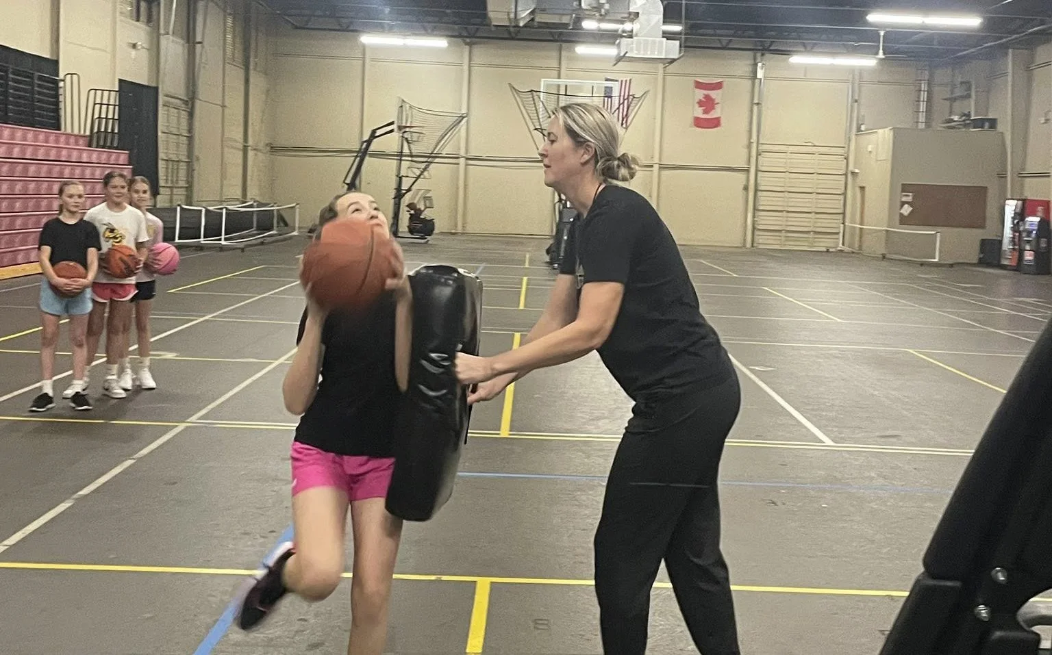A woman coaching a girl in a gymnasium as she practices basketball, with other girls holding basketballs in a line in the background.