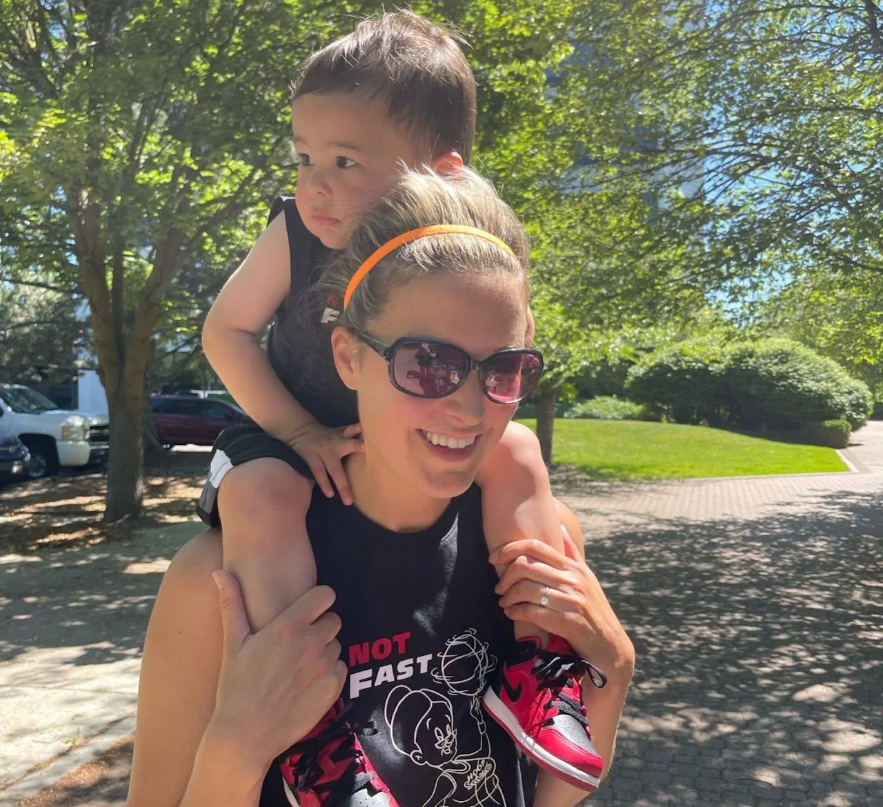 A woman carrying a young boy on her shoulders outdoors on a sunny day, with trees and a park in the background.