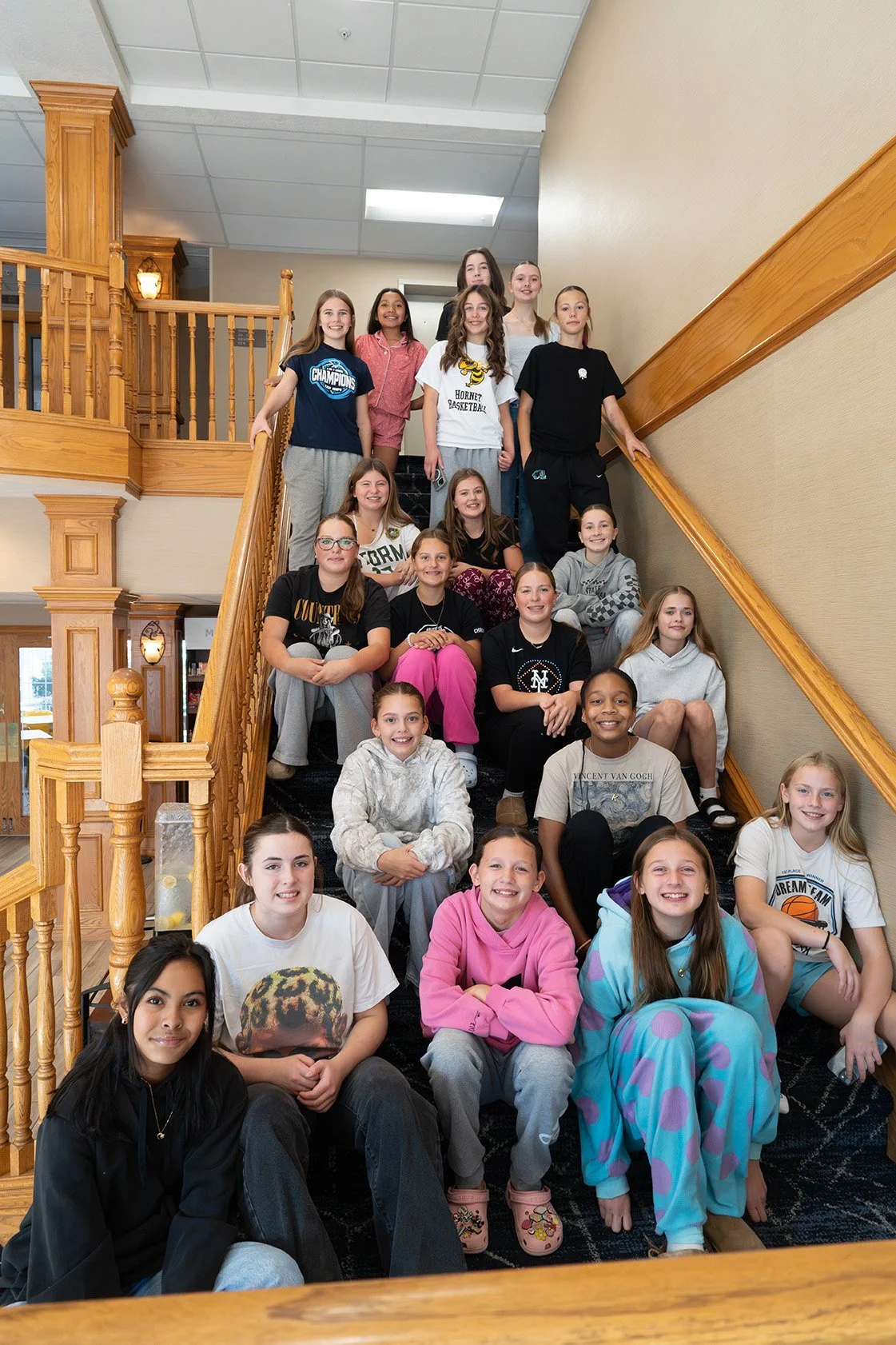 Group of young girls sitting on a staircase in a building with wooden railings and beige walls, smiling at the camera.