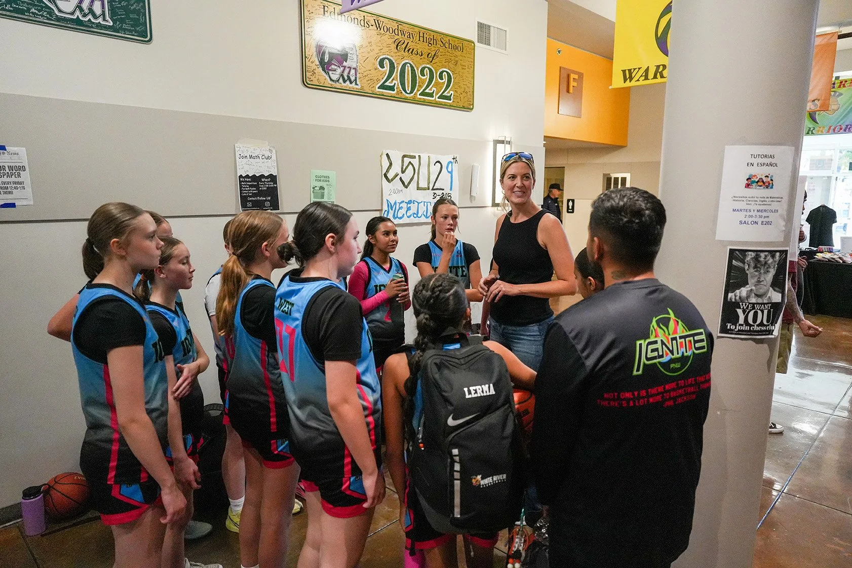 A group of young female basketball players in blue and black uniforms are gathered around their coach, who is speaking to them in an indoor school setting.