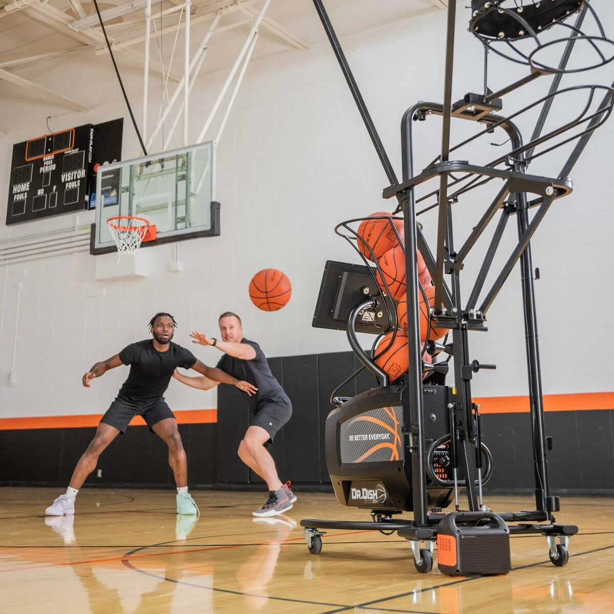 Two men playing basketball in an indoor gym, with a basketball shooting machine dispensing basketballs nearby.