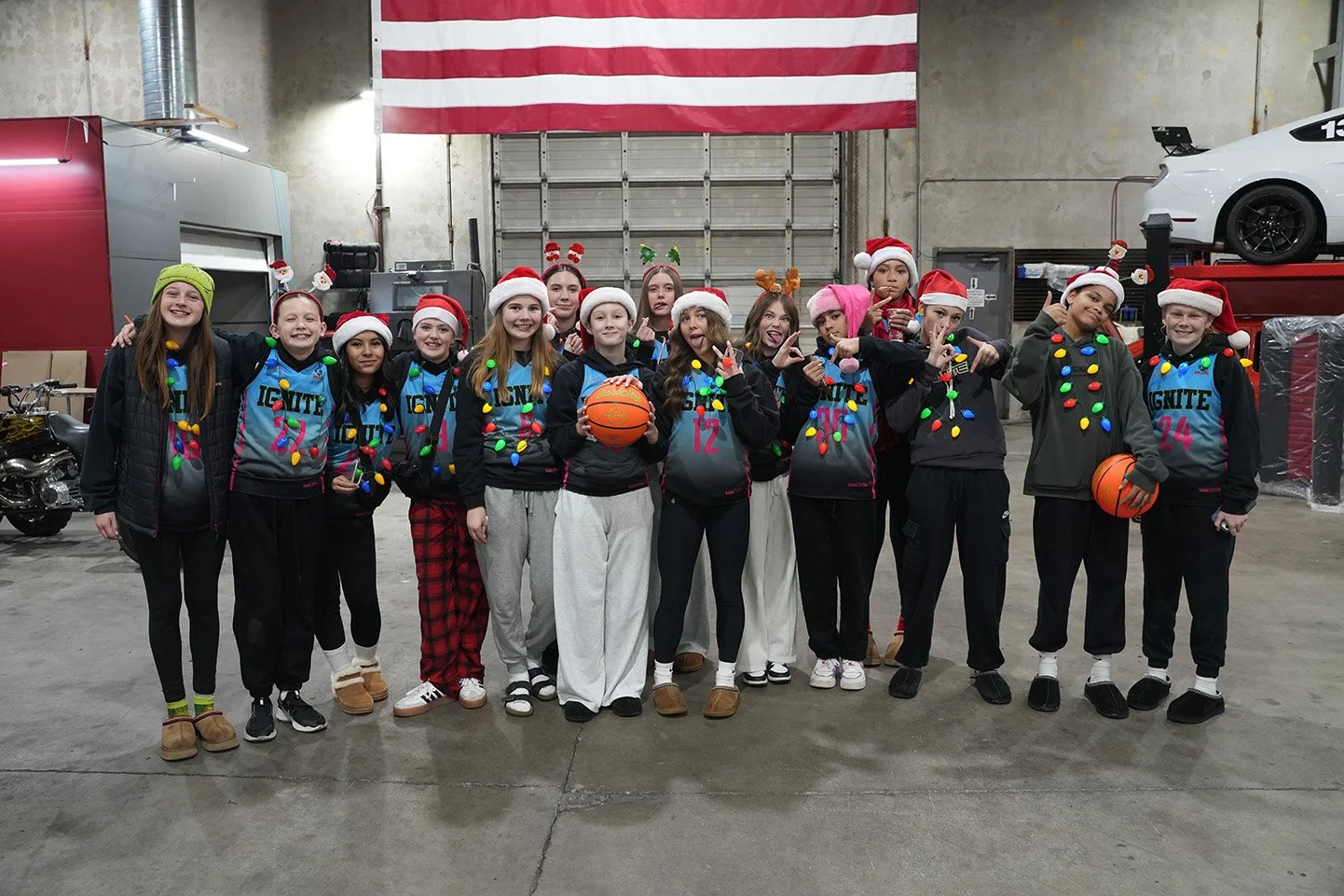 Group of teenagers celebrating Christmas with festive hats and ornaments, holding basketballs, in an indoor garage with an American flag in the background.