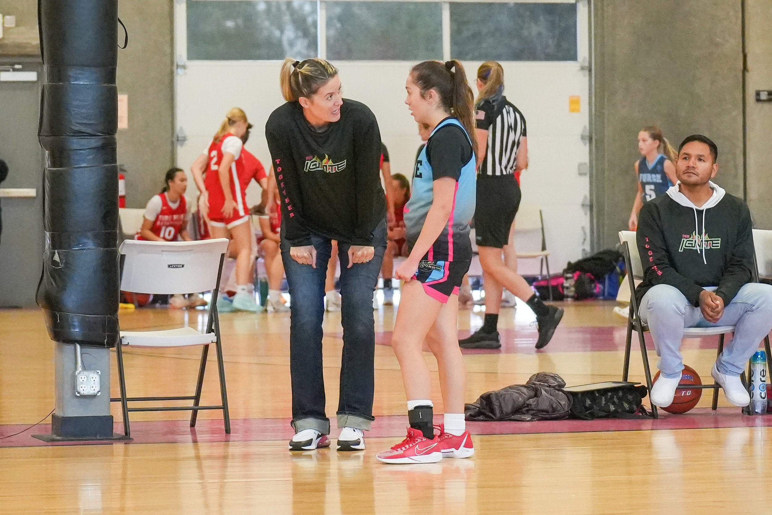 A female basketball player in a black, pink, and blue uniform stands on the court, listening to her coach who is leaning forward and speaking to her. Other players and a referee are visible in the background.