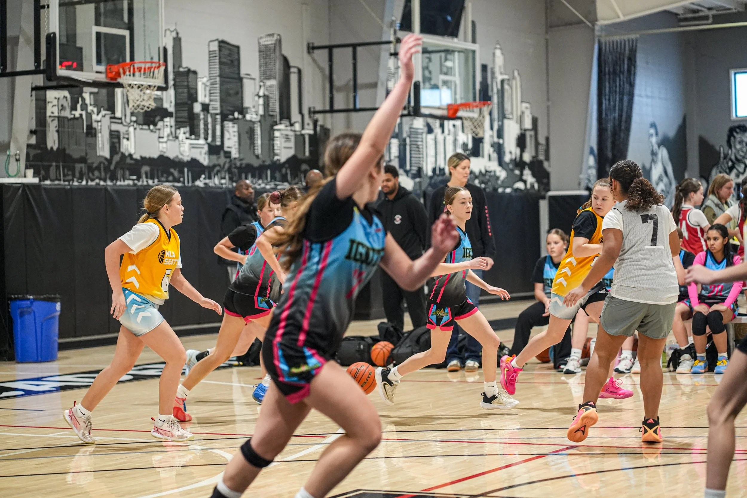 Girls playing basketball on indoor court with urban cityscape mural in background.