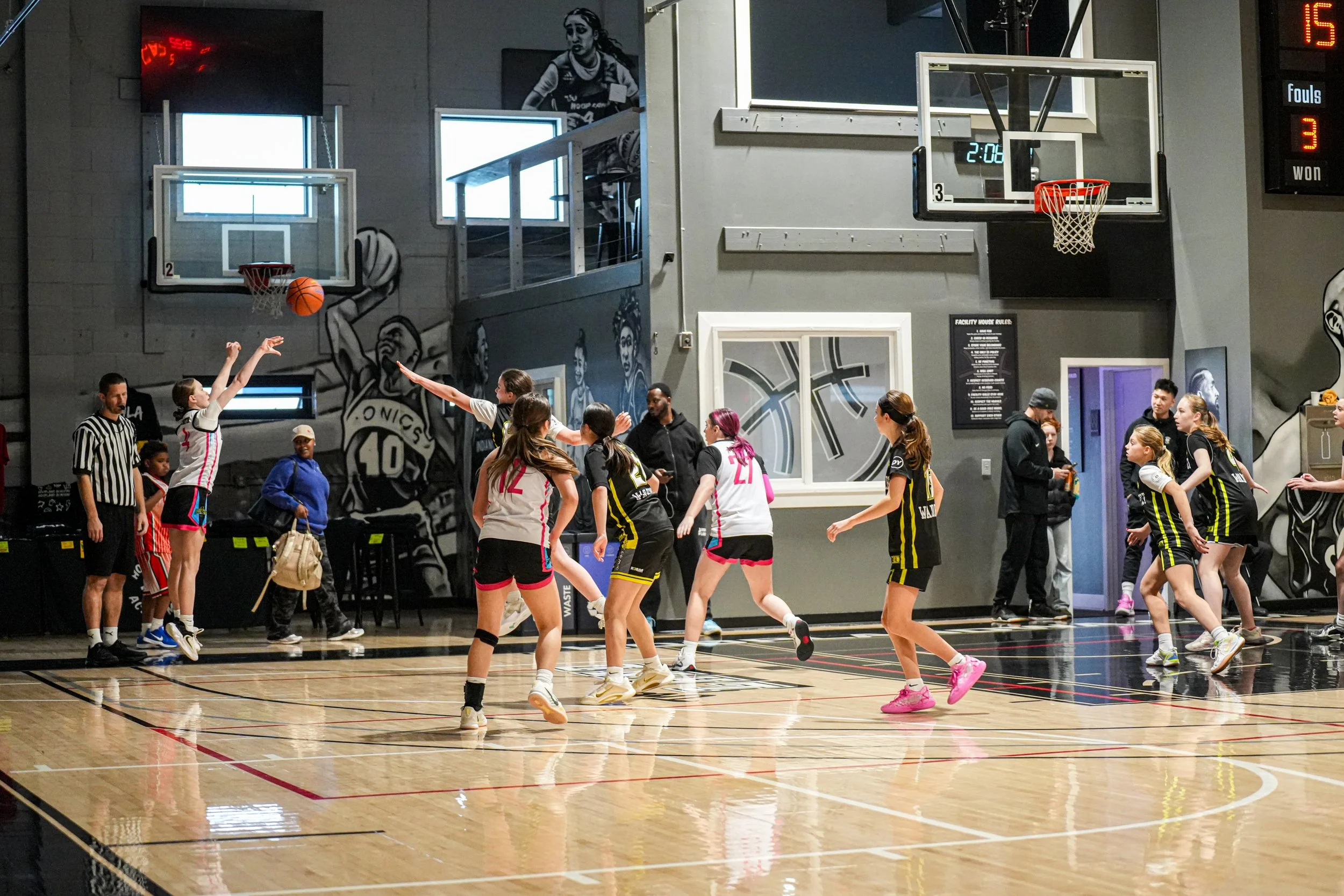 An indoor basketball game with both teams playing, with some players jumping and reaching for the ball. The court has a wooden floor, and there are referees and spectators visible in the background.