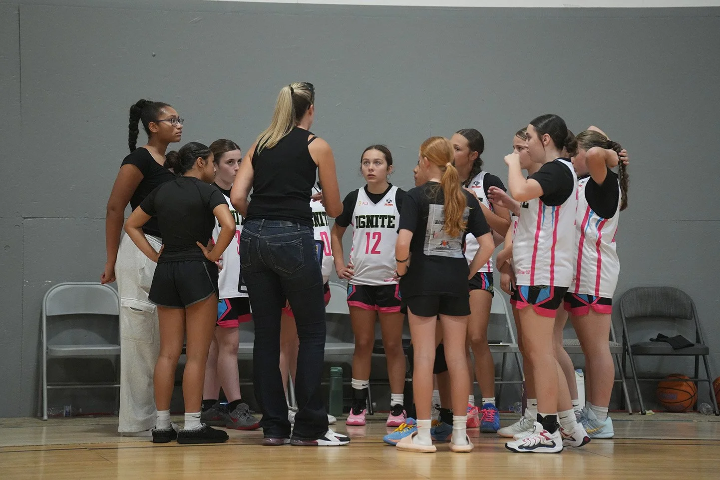 A group of female basketball players in uniforms gathered around their coach during a timeout on the court.
