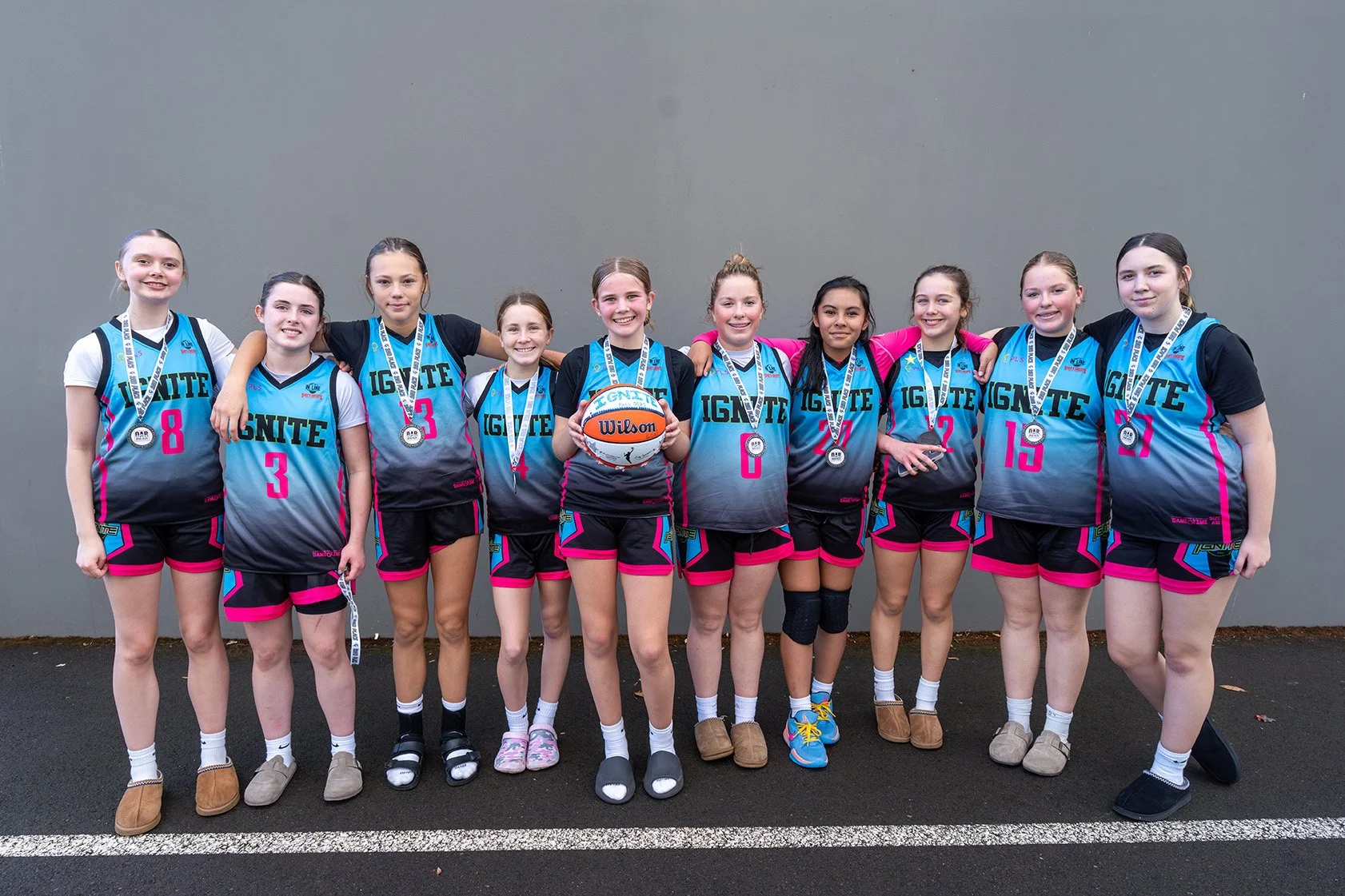 Girls' basketball team wearing matching uniforms with medals around necks, some holding a basketball, standing on a paved surface against a gray wall.