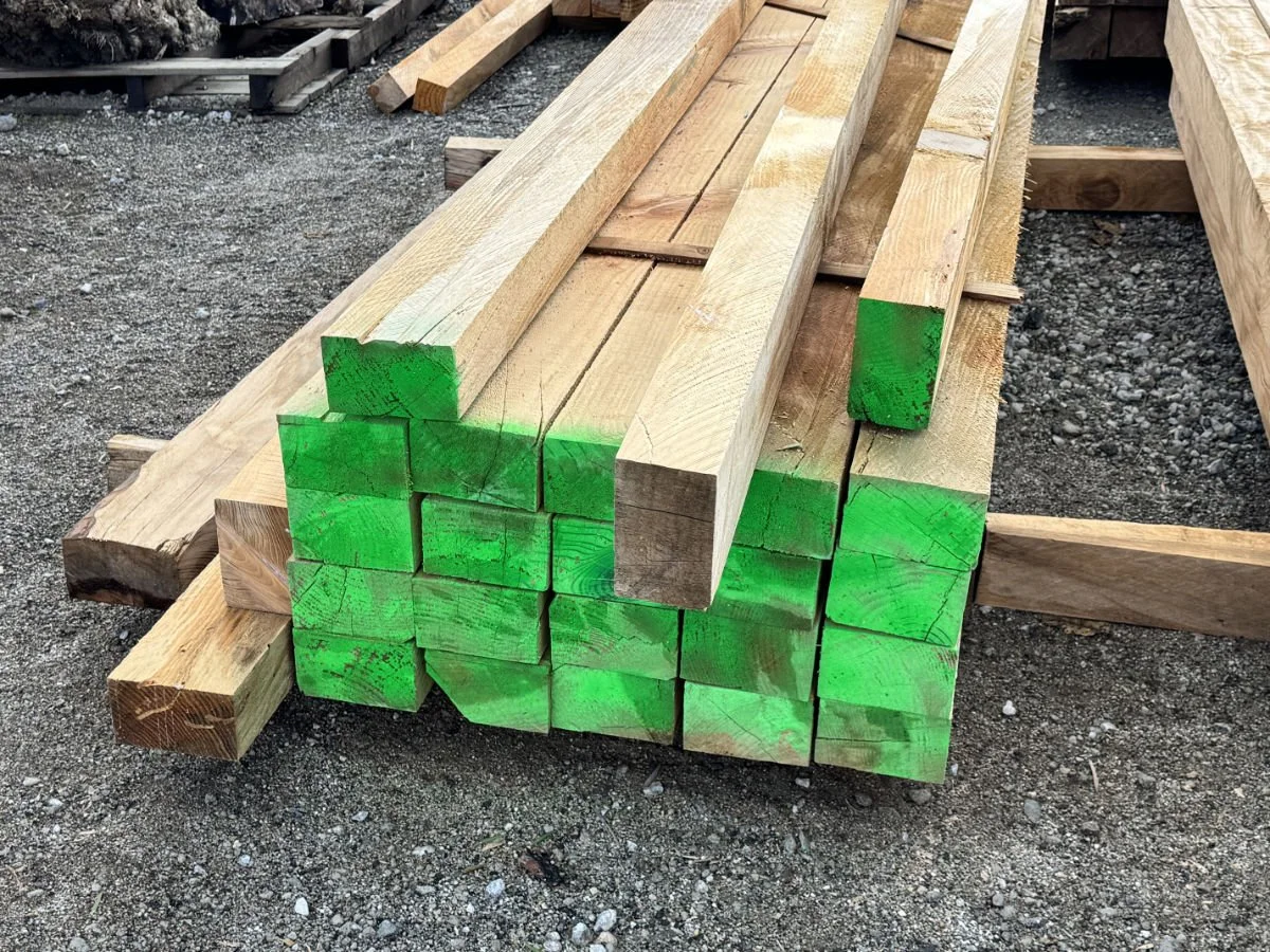 Stacked wooden planks on gravel ground, with some ends painted green, at a construction site.