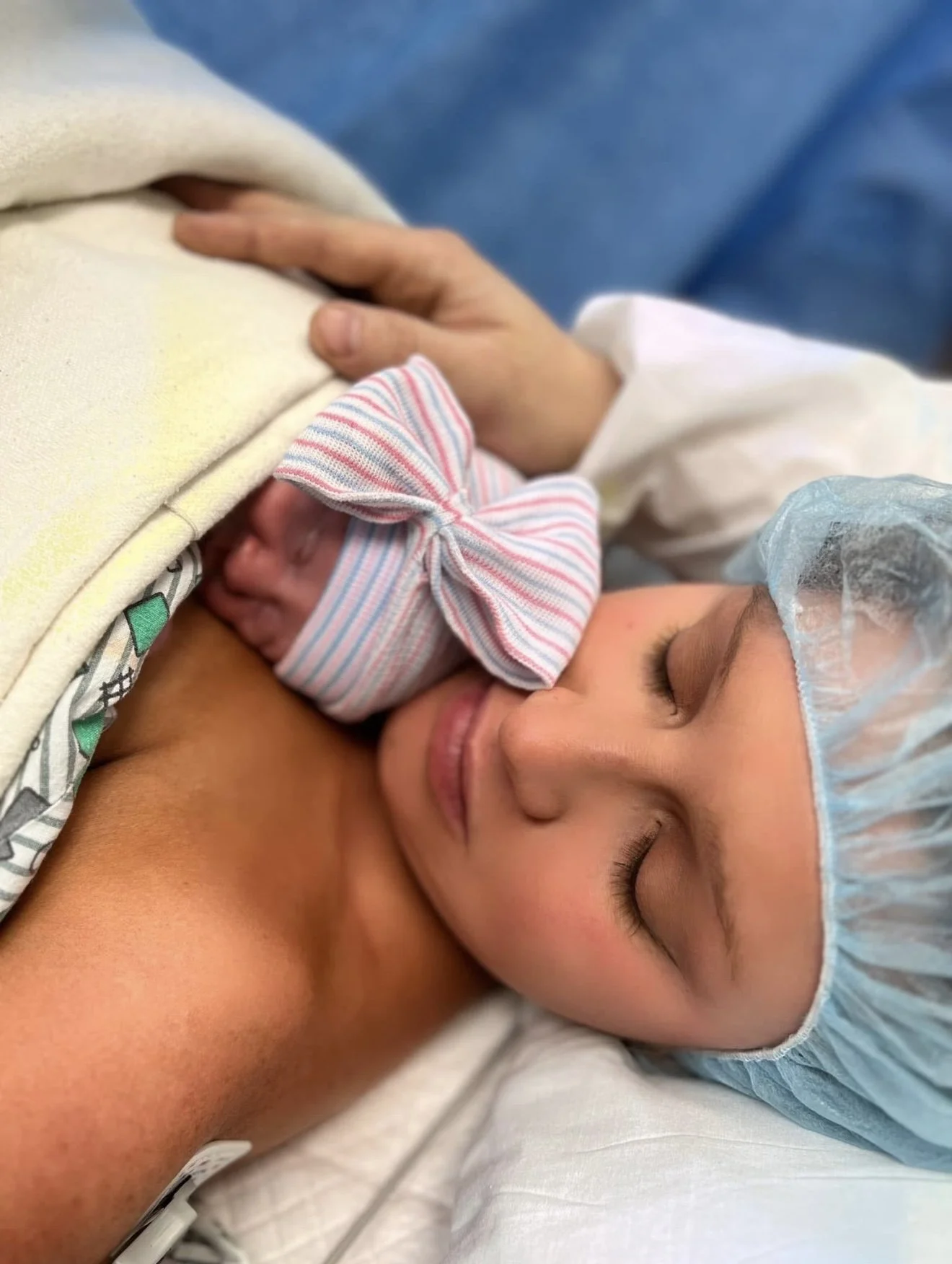 A woman, wearing a hospital cap, is holding a newborn baby close to her chest after childbirth, both with their eyes closed.