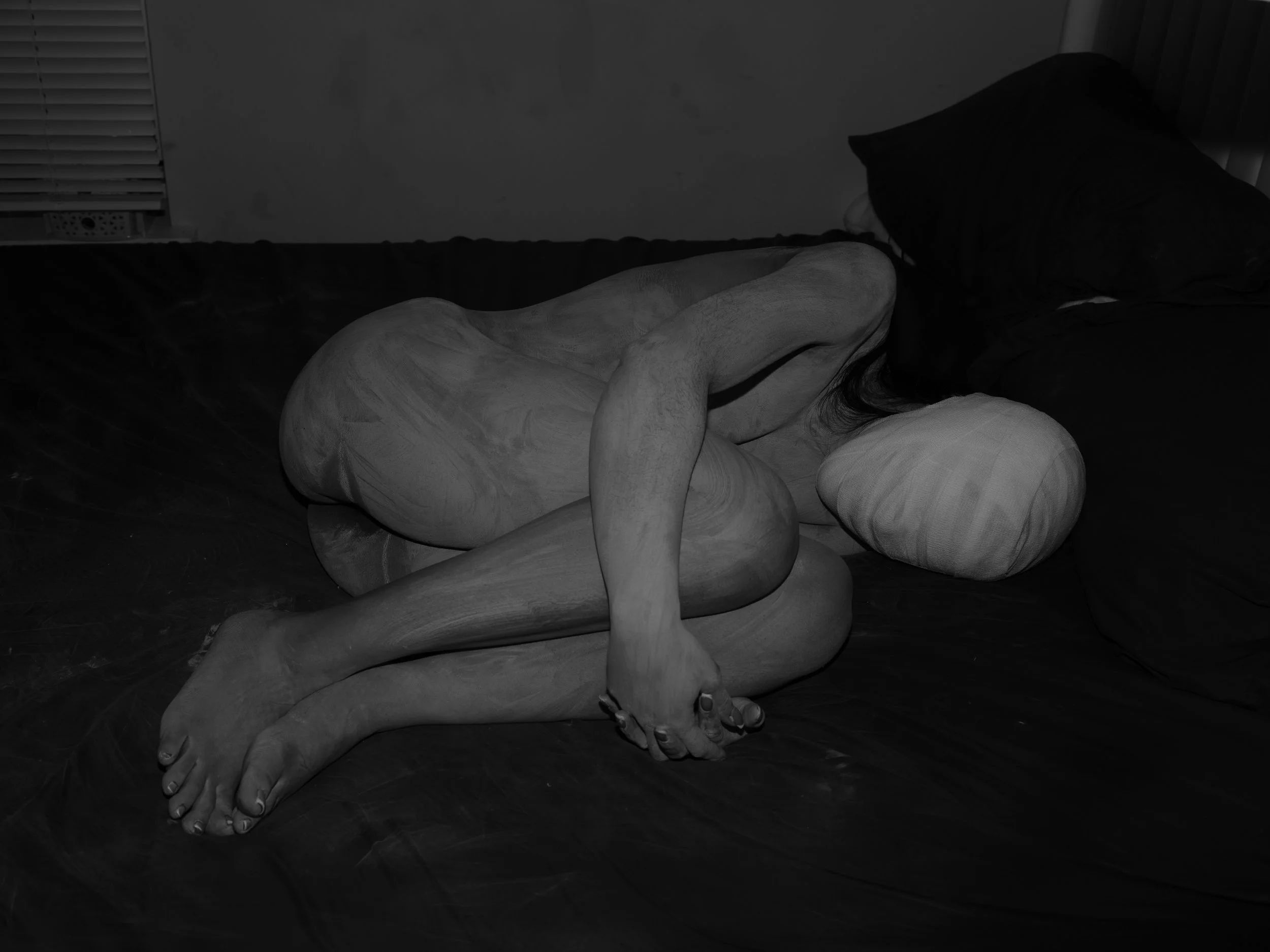 A person lying on a bed with their body curled into a fetal position, with their face turned away and covered by a black cloth or pillow, in black and white.