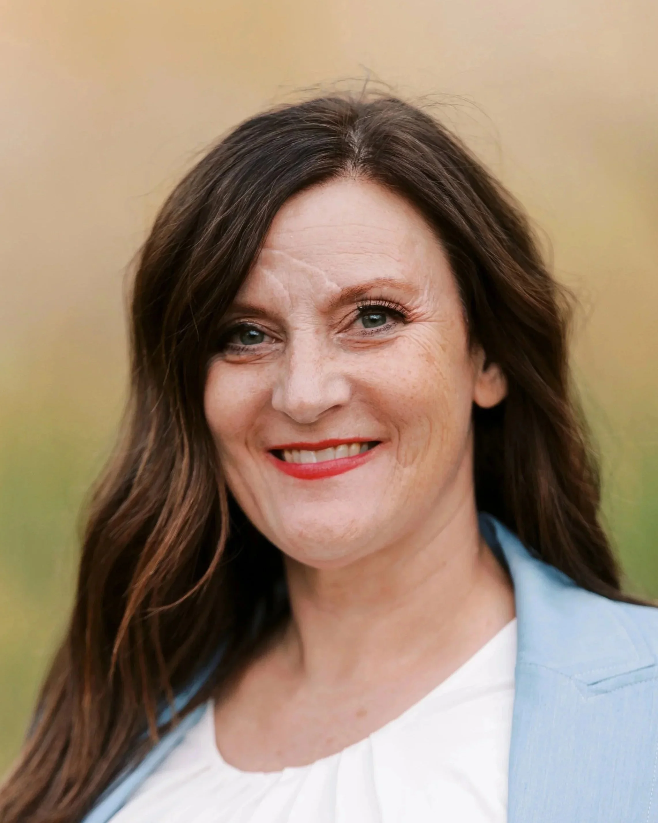 A woman with long brown hair and blue eyes, smiling outdoors with a blurred natural background.