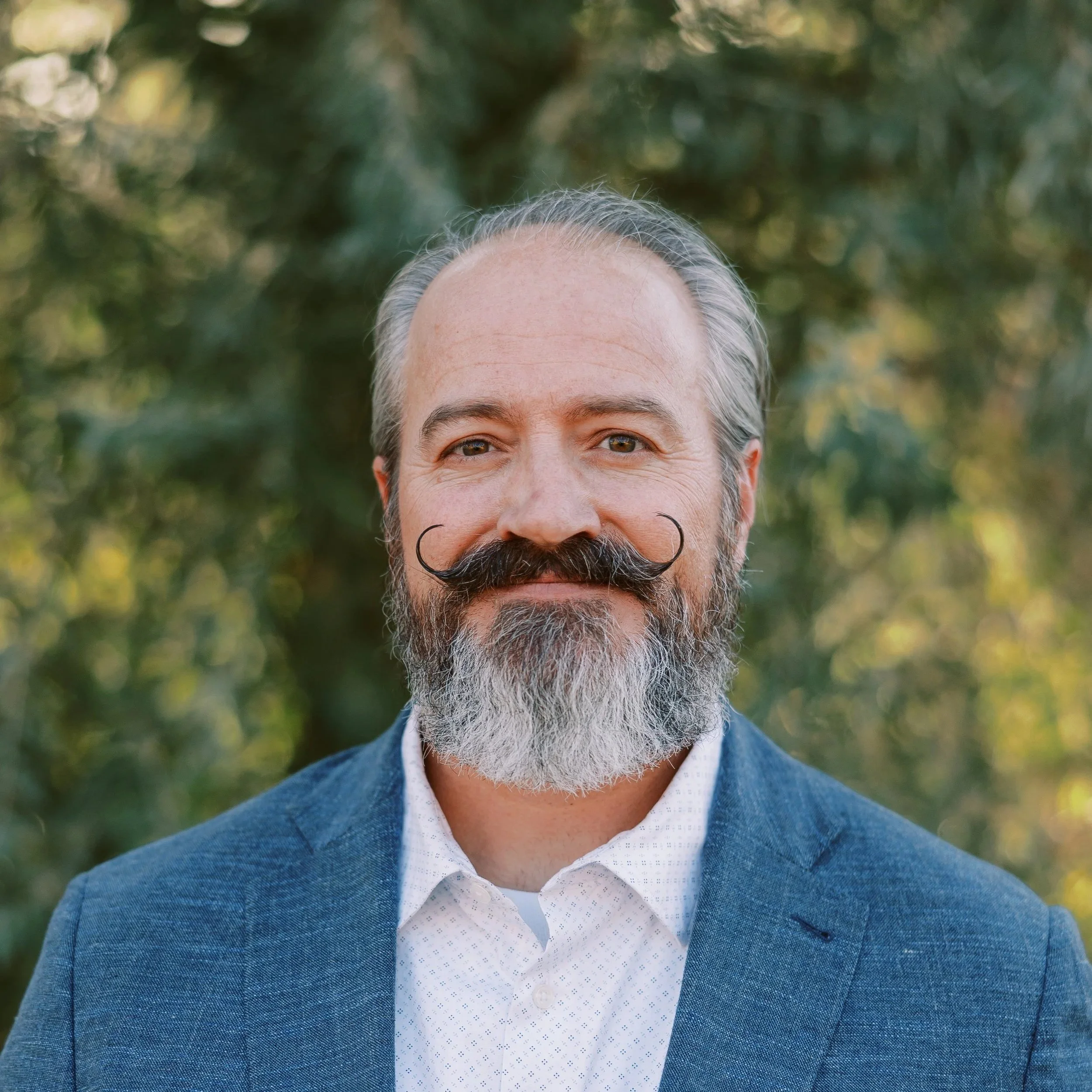A middle-aged man with a salt-and-pepper beard and mustache, smiling, wearing a blue blazer over a white shirt, standing outdoors with blurred green trees in the background.