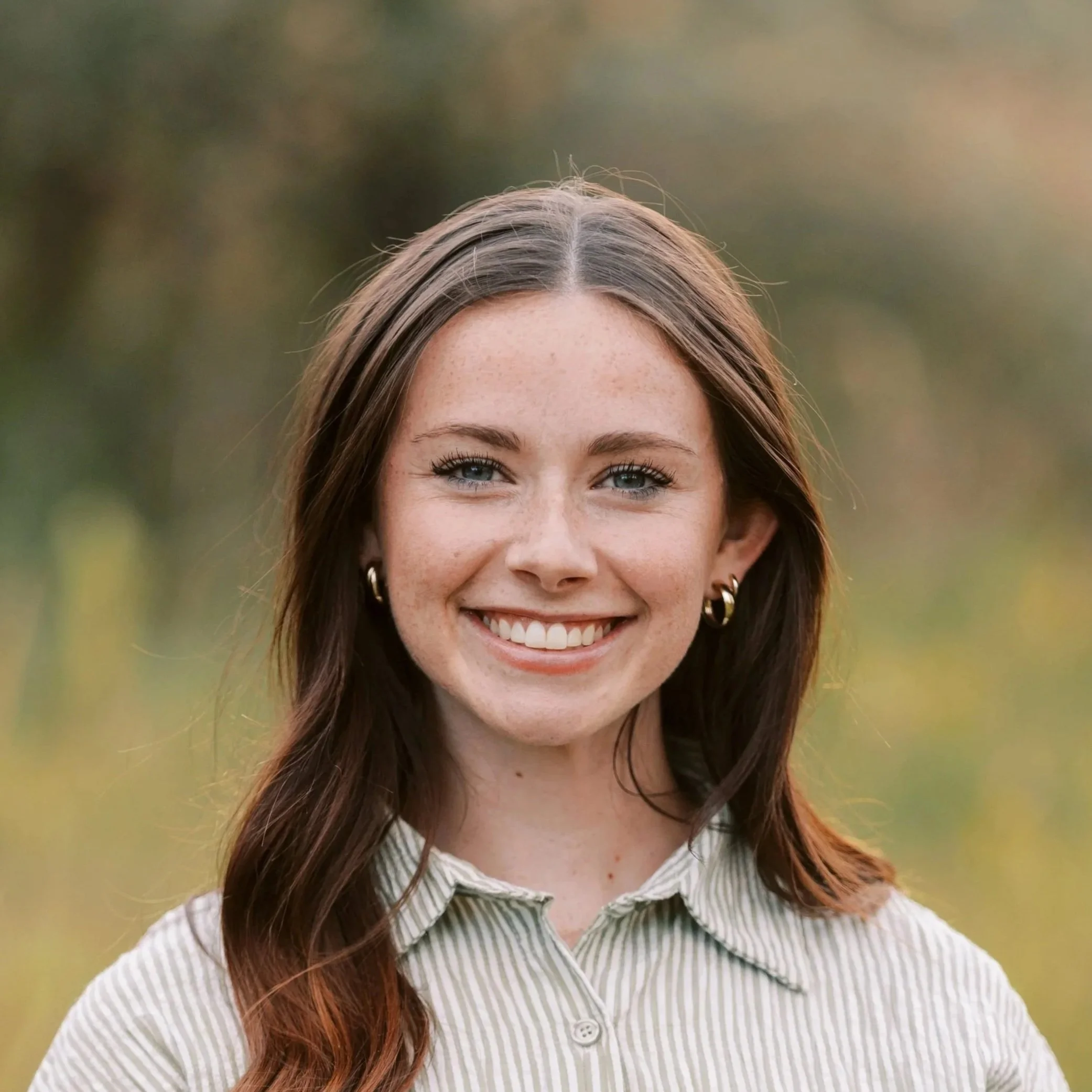 A young woman with long brown hair, blue eyes, and freckles smiling outdoors in a natural setting with soft-focus greenery in the background.