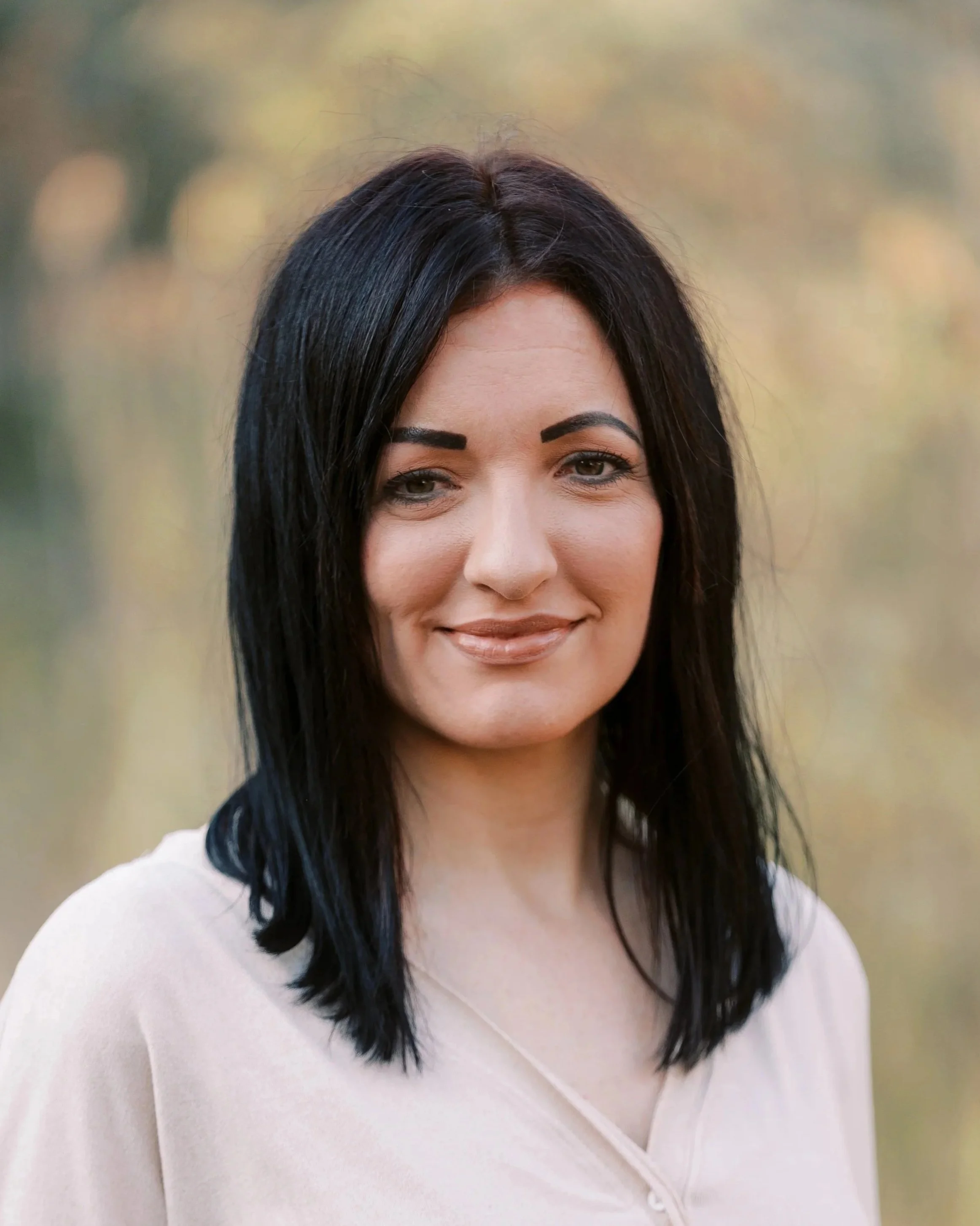 A portrait of a woman with black hair wearing a light-colored top, standing outdoors with a blurred background of trees and foliage.