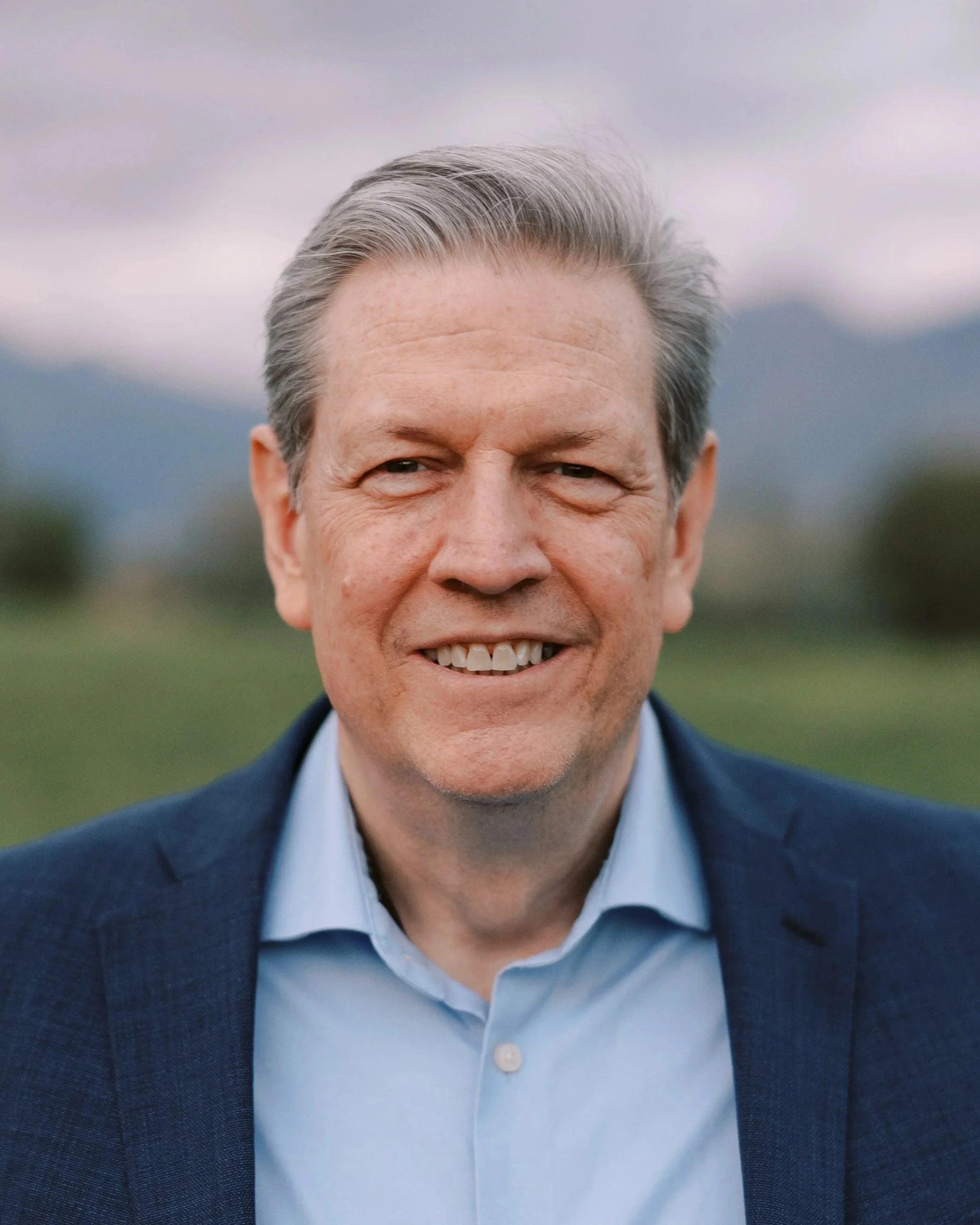 Close-up image of a smiling middle-aged man with gray hair, wearing a blue shirt and navy blazer, outside with blurred green landscape and cloudy sky in the background.
