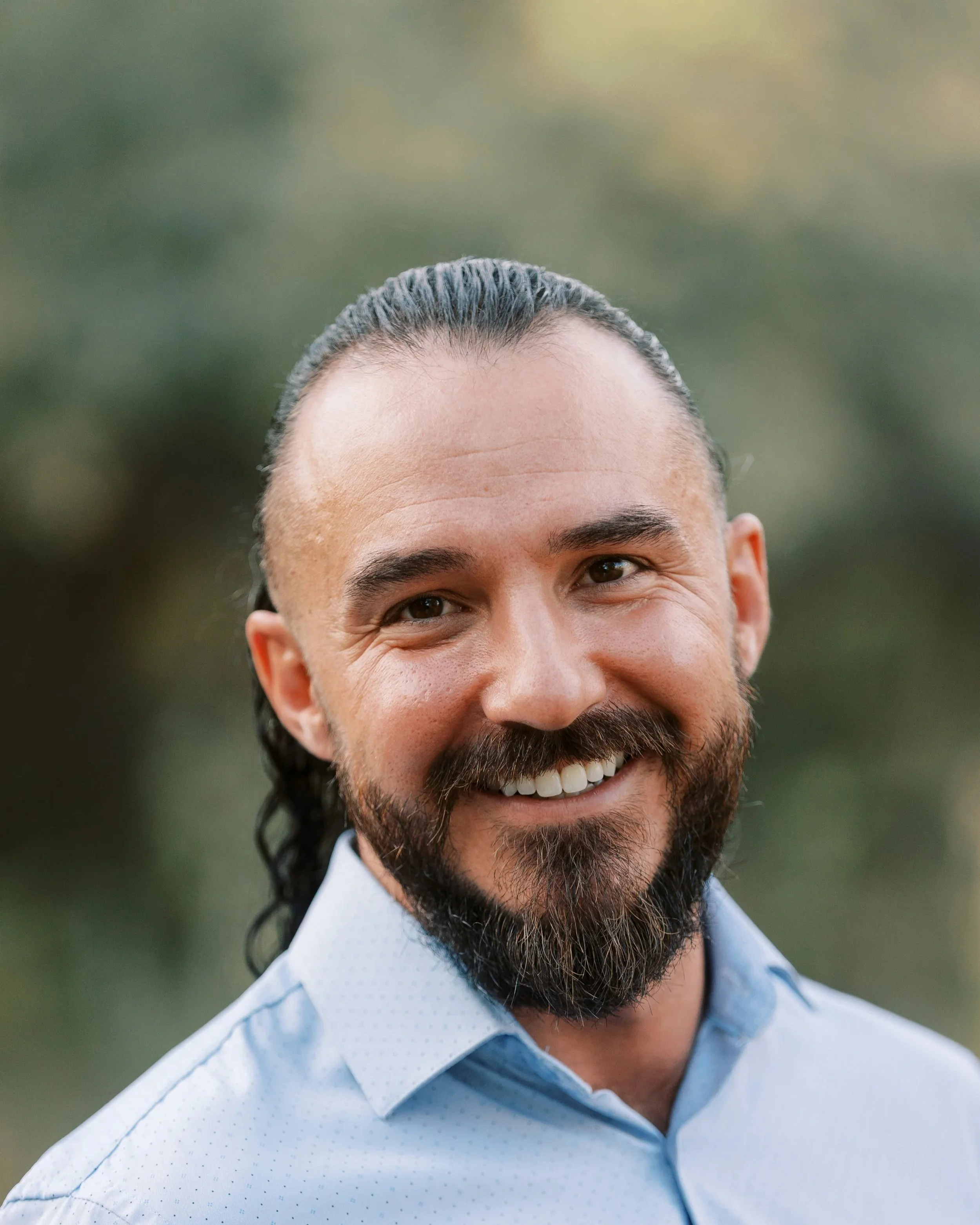 Close-up of a smiling man with a dark beard and long dark hair tied back, wearing a light blue collared shirt, outdoors with blurred greenery in the background.
