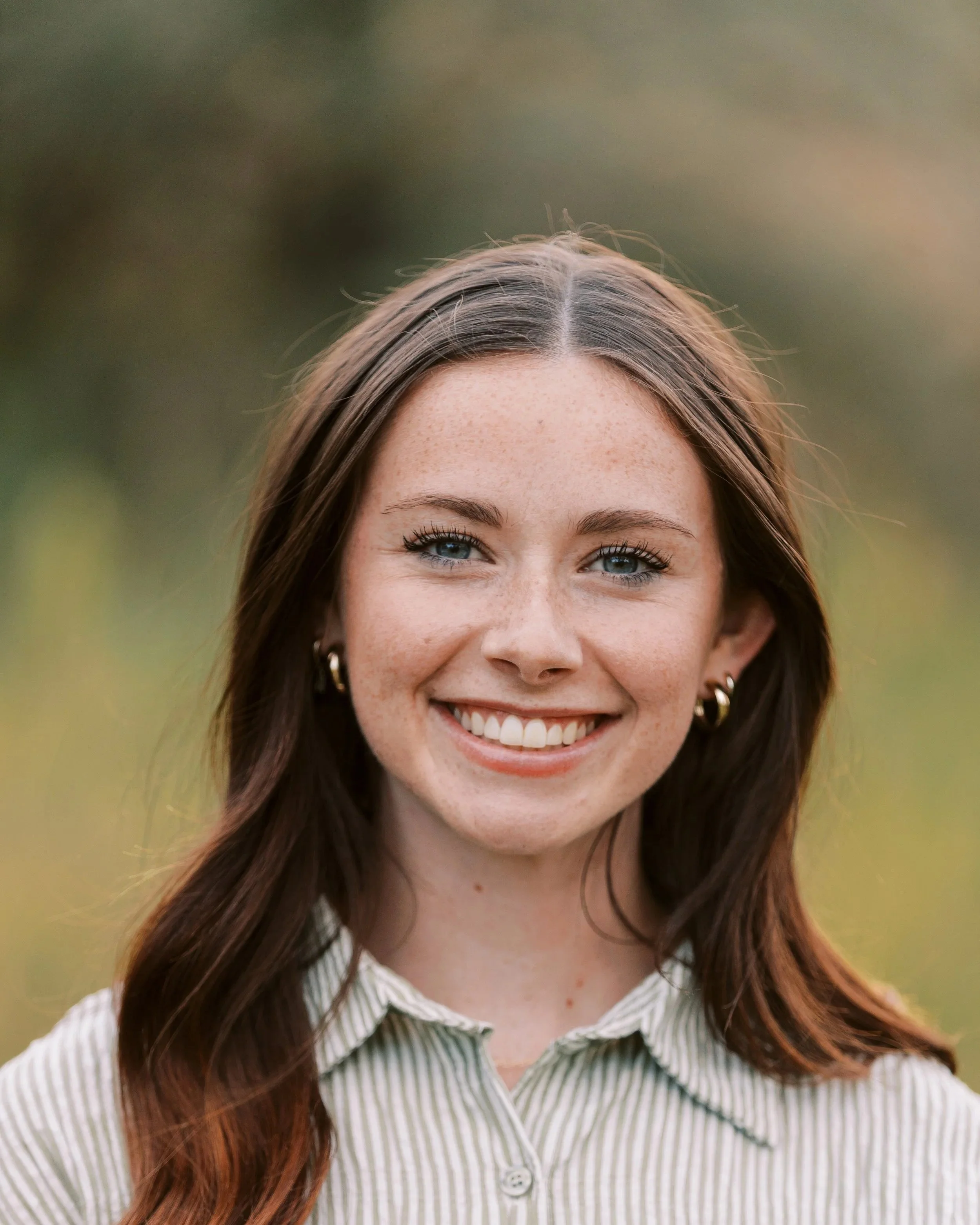 Close-up portrait of a young woman with brown hair, blue eyes, and freckles, smiling outdoors with a blurred natural background.