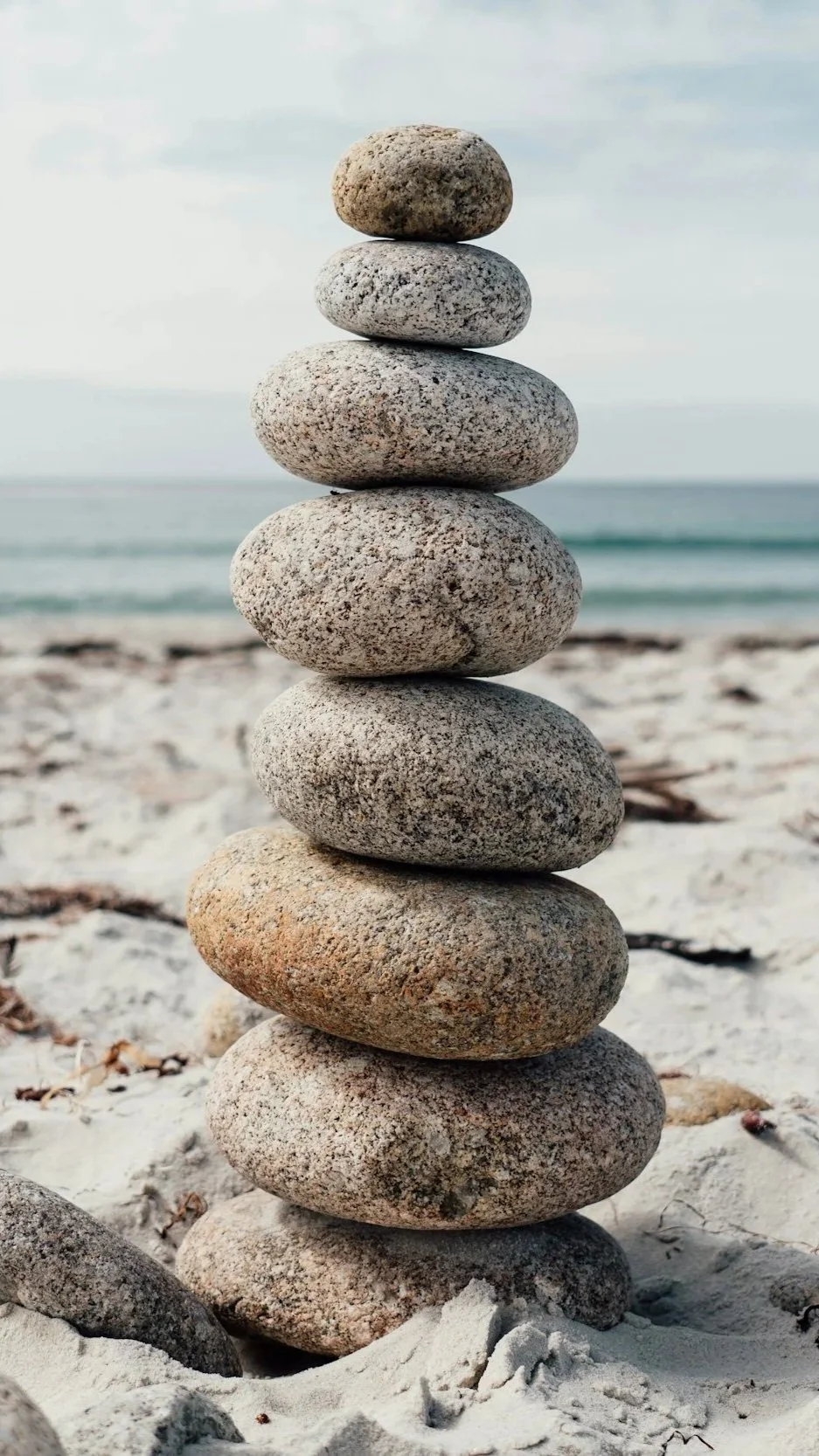 A stack of seven smooth stones balanced on a sandy beach with the ocean and sky in the background.