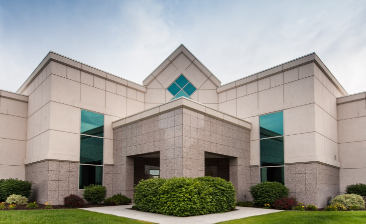 Front view of a modern office building with beige and gray exterior walls, green bushes, and large tinted windows.