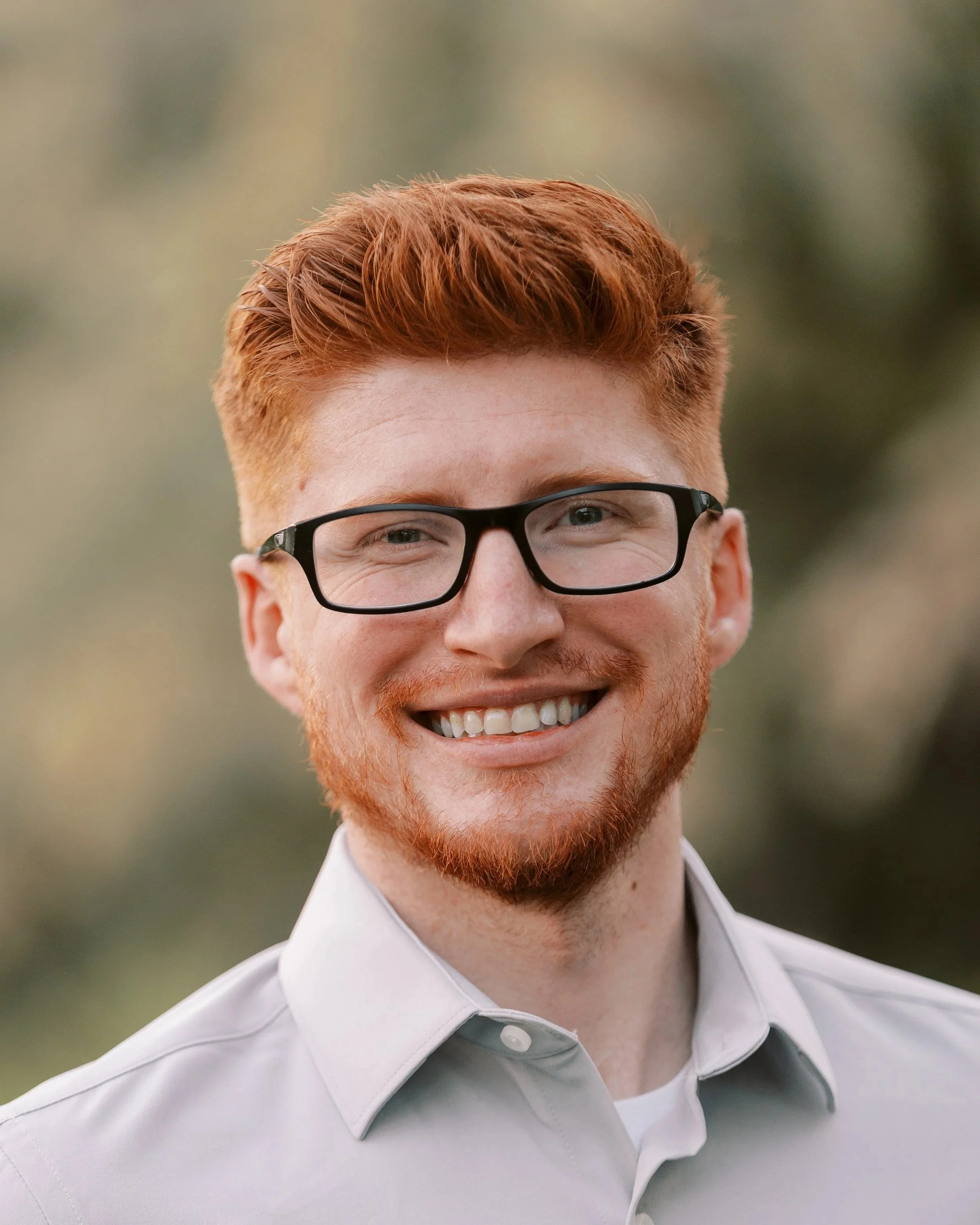 A smiling man with red hair, beard, glasses, and wearing a light gray shirt outdoors.