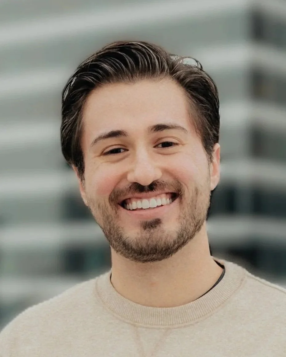 Portrait of a smiling man with dark hair, beard, and a beige sweater, standing outdoors with a blurred cityscape background.