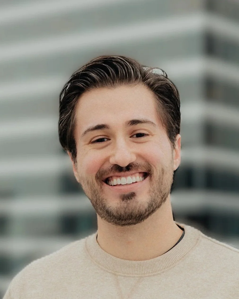 Close-up of a smiling young man with dark hair and a beard, wearing a beige sweater, blurry outdoor background.