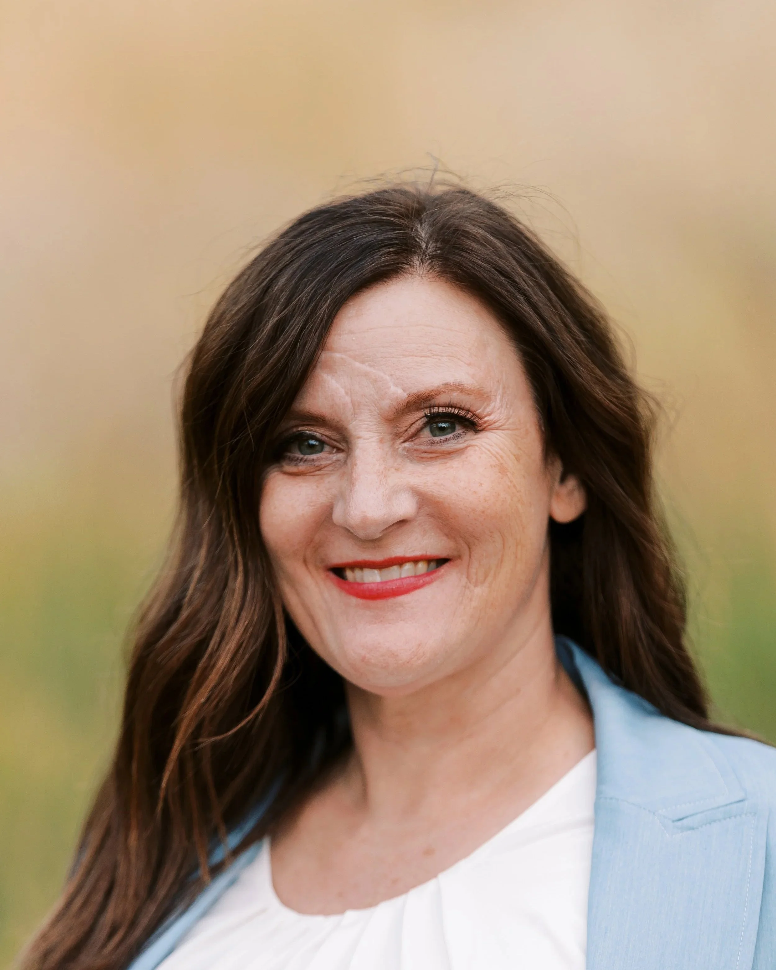 A smiling woman with shoulder-length dark brown hair and red lipstick, wearing a light blue blazer and white blouse, outdoors with a blurred green and yellow background.