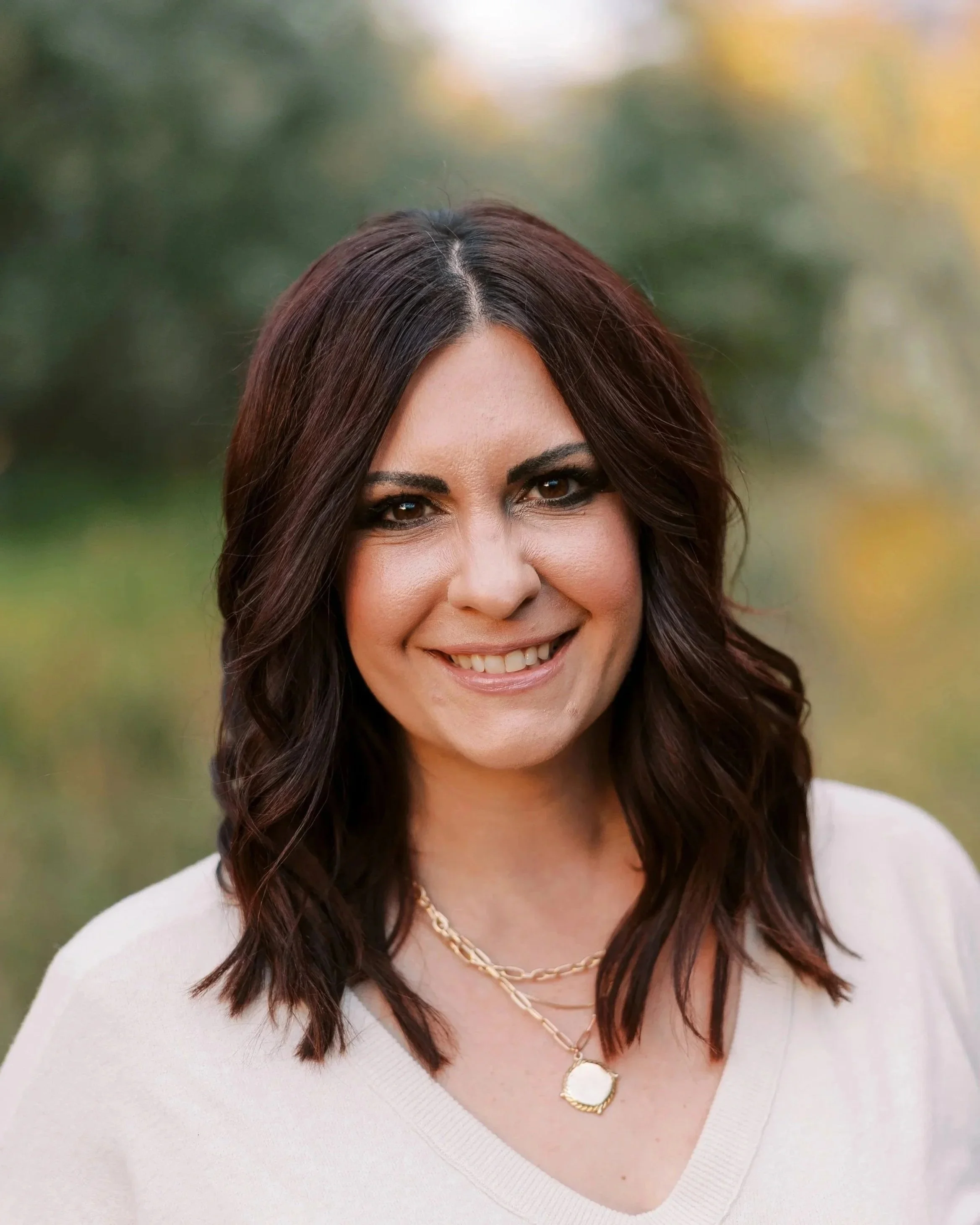 Headshot of a woman with shoulder-length dark brown hair, smiling, wearing a white top and layered gold necklaces, outdoors with blurred greenery background.